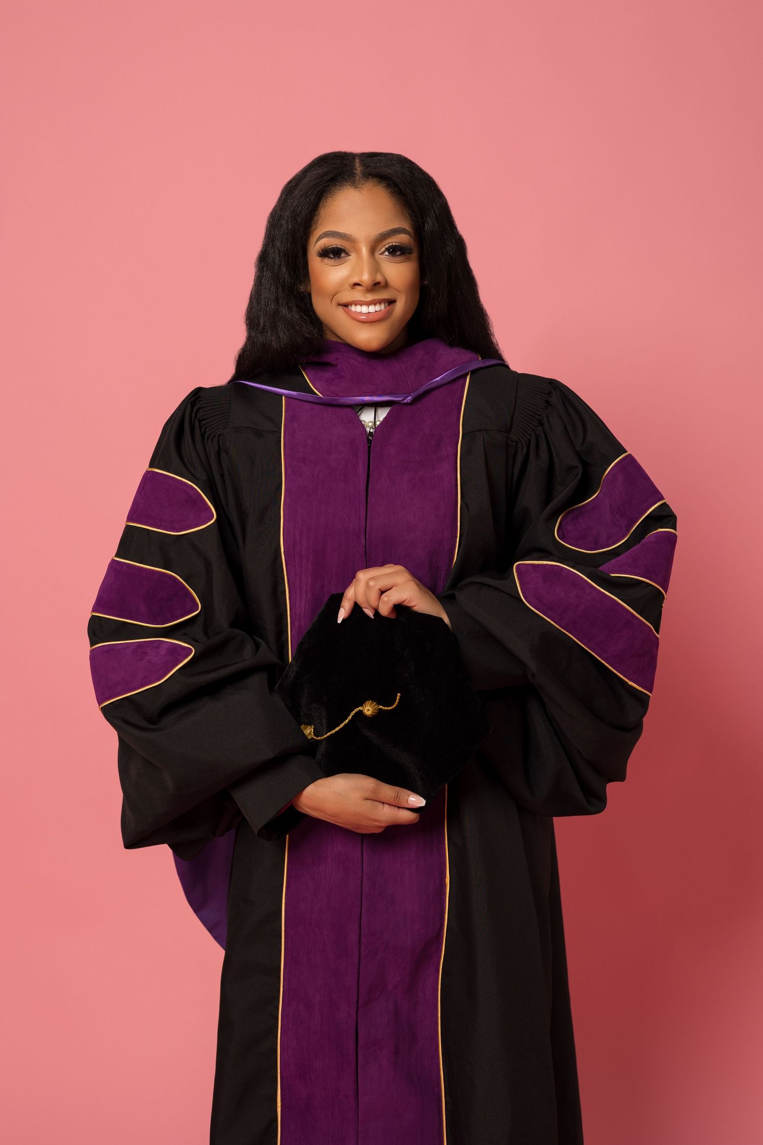 Academic regalia featuring black and purple doctoral gown against pink background.