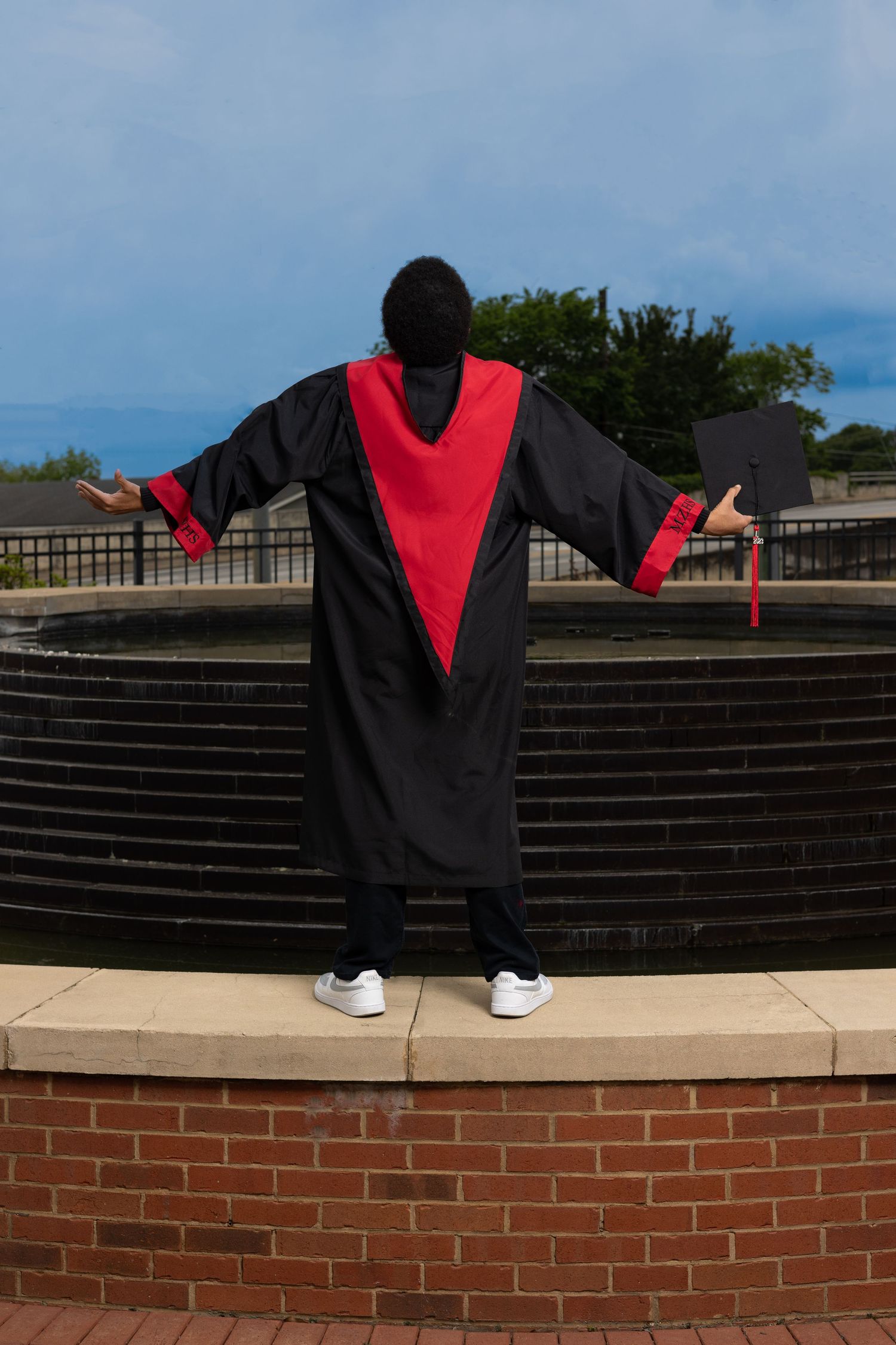 Graduate in black and red regalia stands with arms outstretched on brick wall.