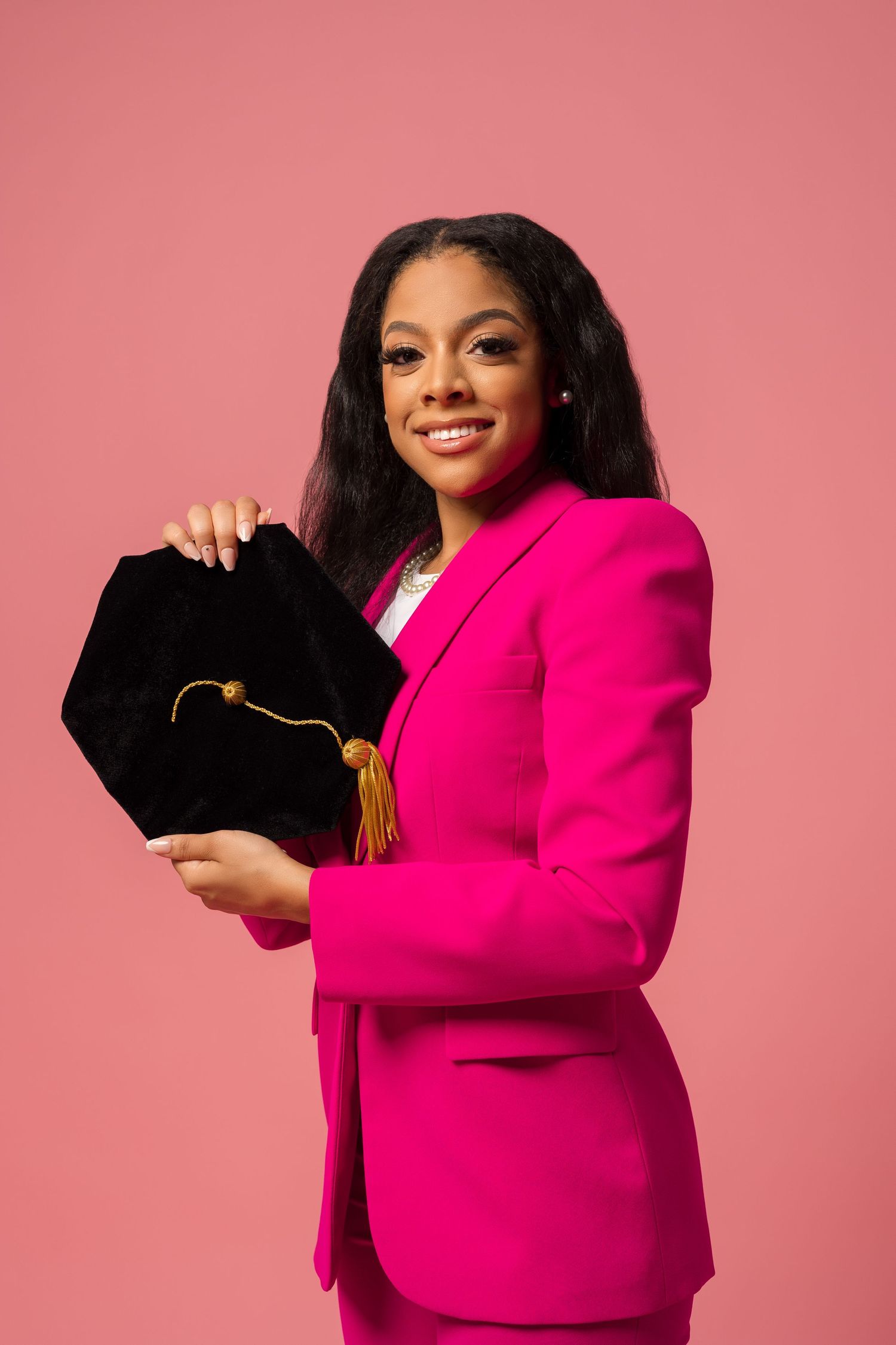 Professional wearing bright pink suit holding black graduation tassel bag against pink backdrop.