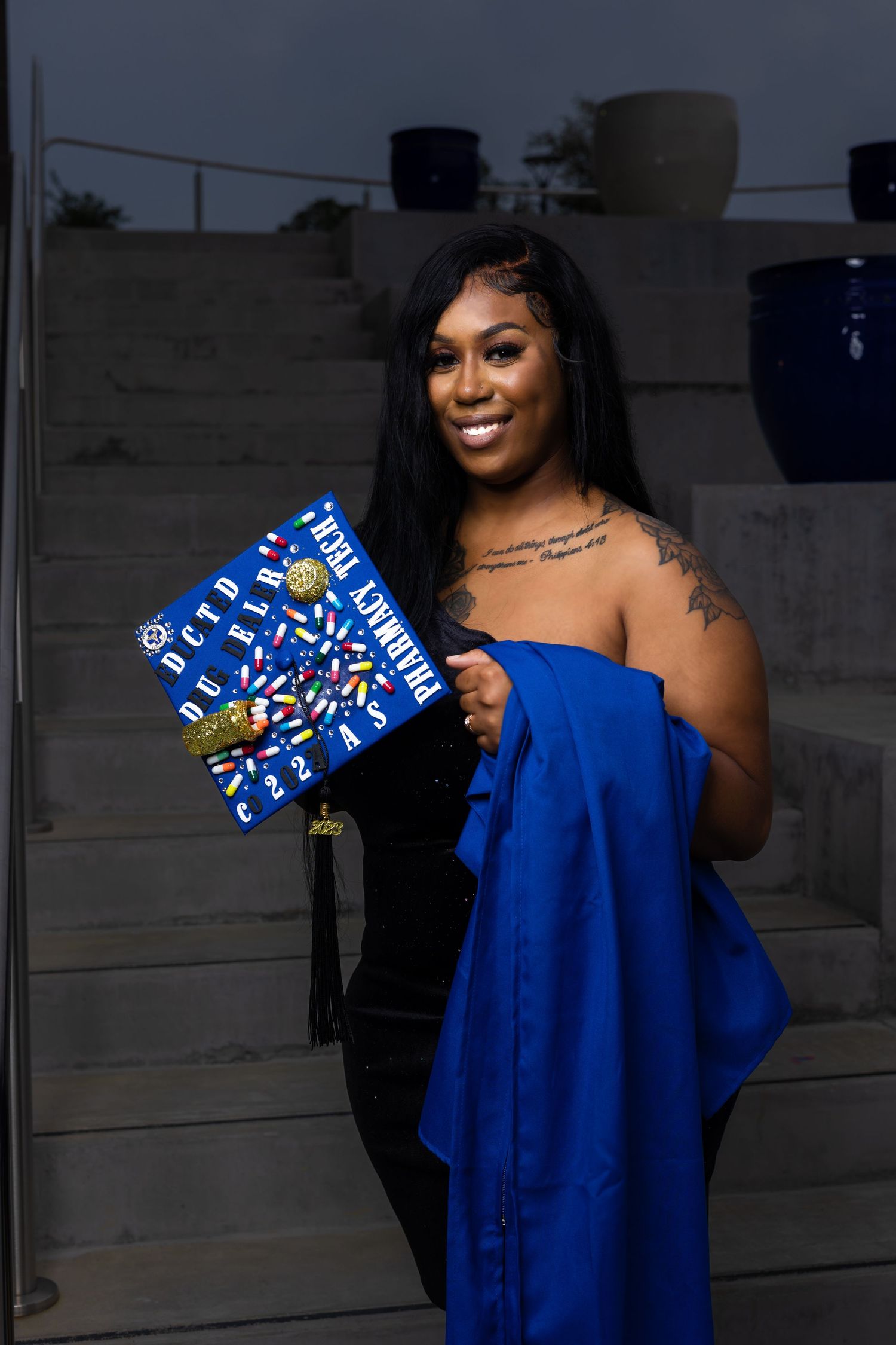 Graduate holding decorated cap while wearing blue graduation attire at night.