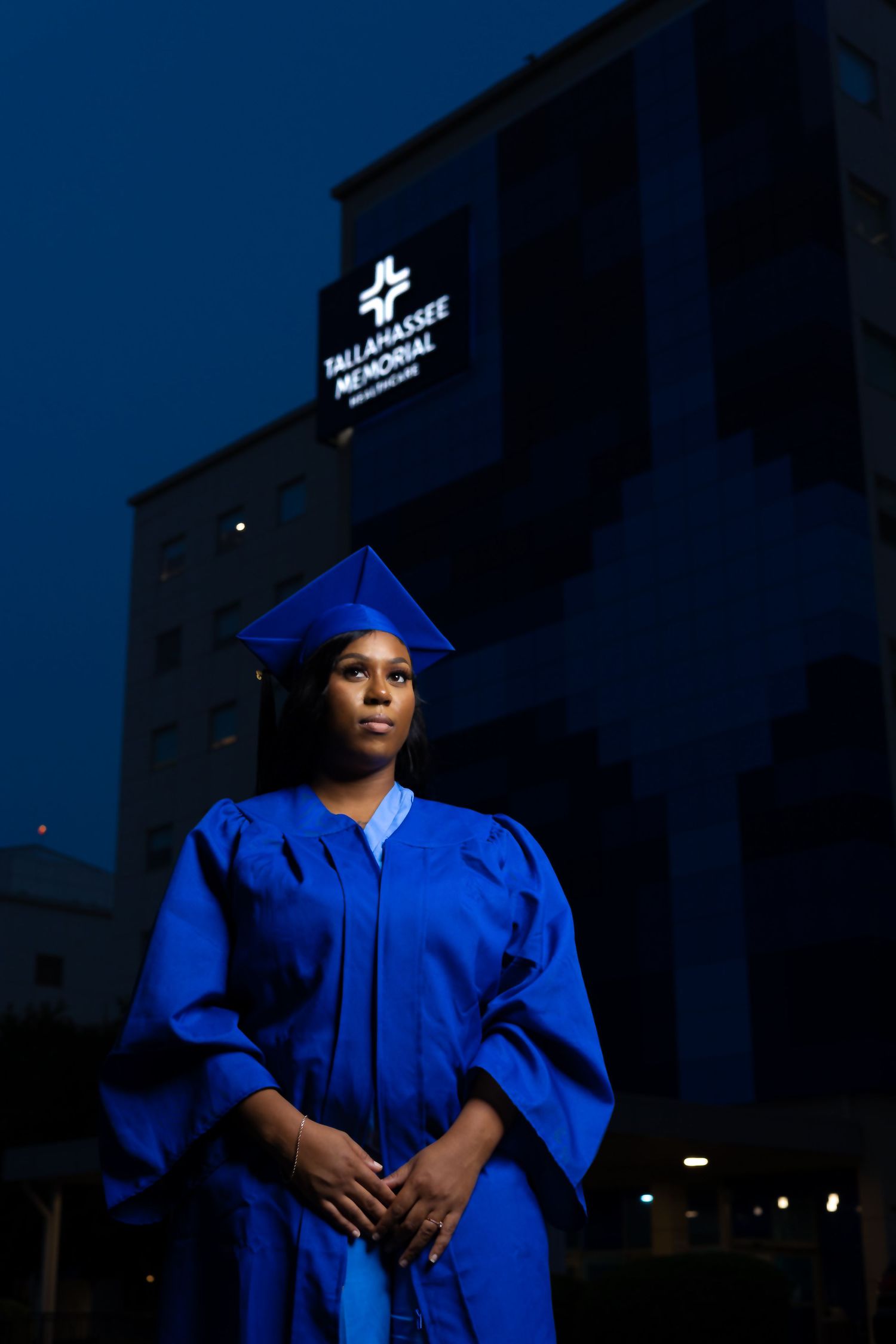 Graduate in blue regalia standing in front of illuminated hospital sign at night.