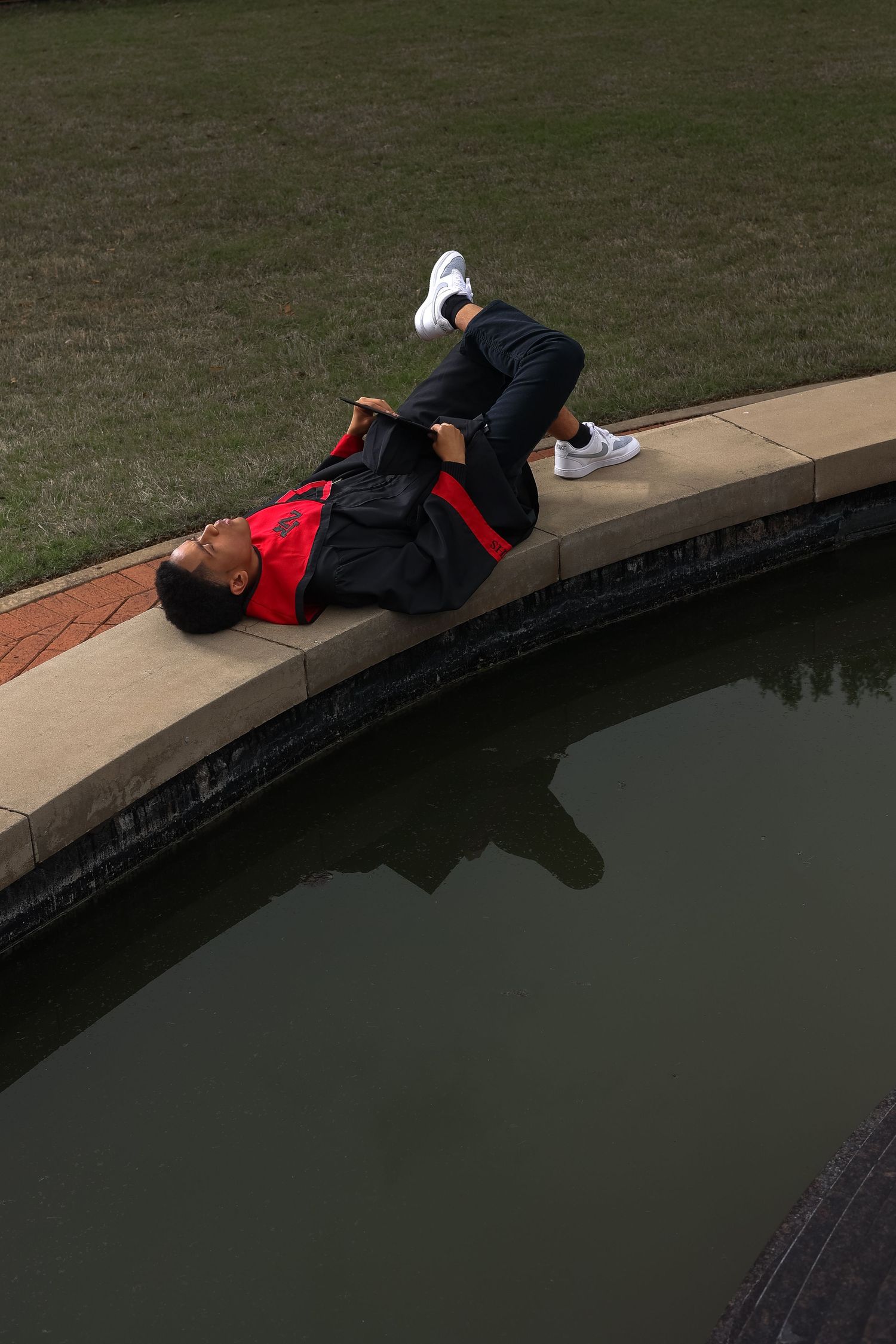Graduate in academic regalia lies on edge of water feature for creative photo.