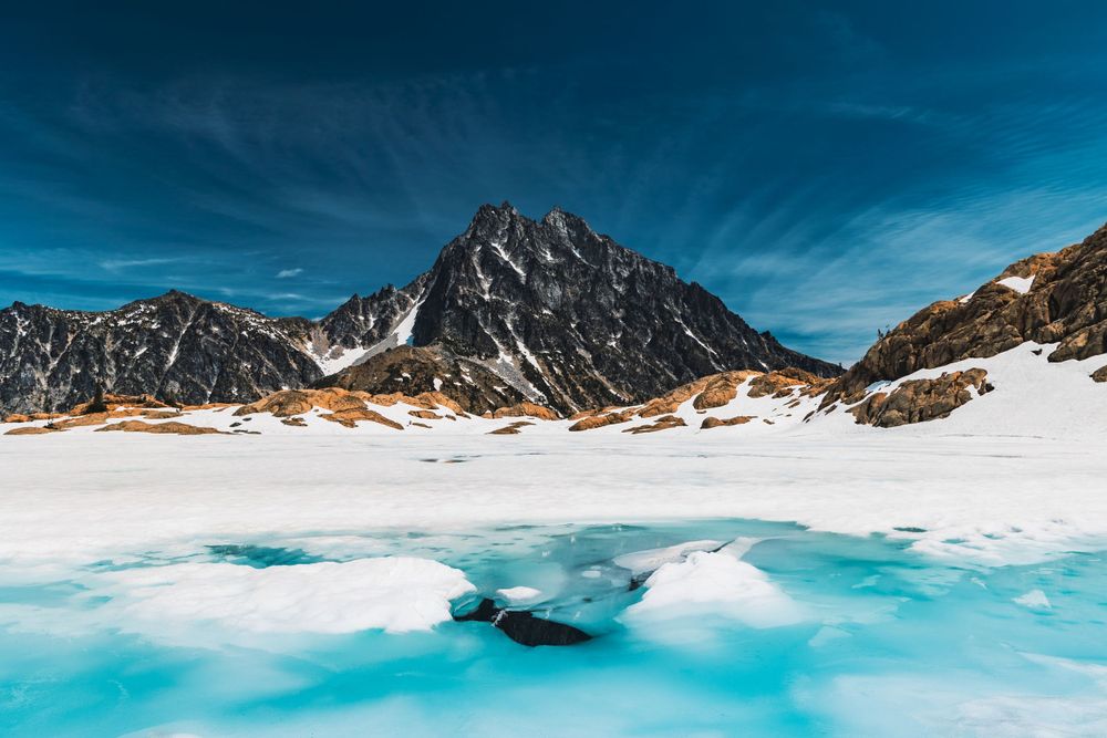 Snow-covered mountain peak with turquoise glacial lake in the foreground on a sunny winter day.