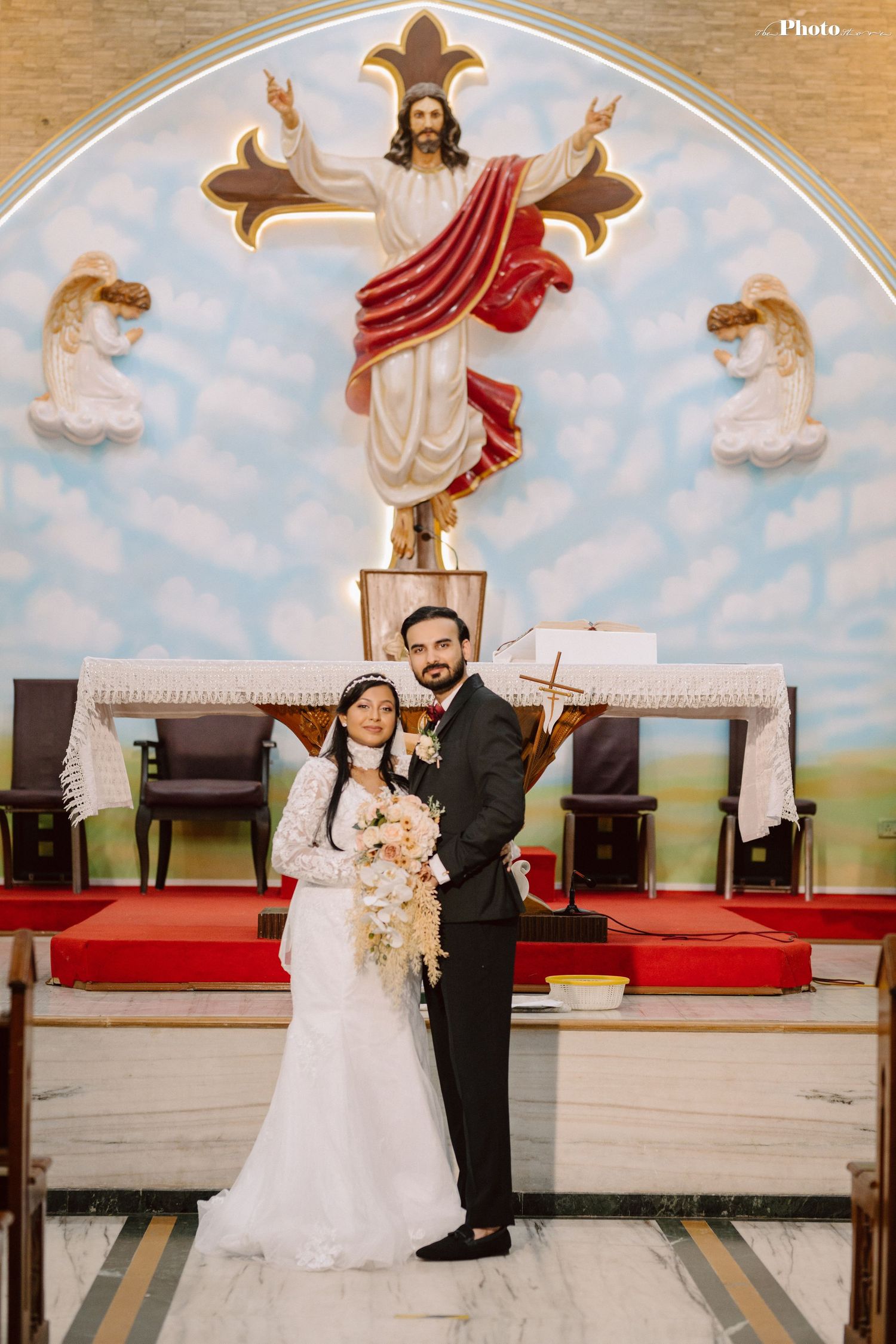 Wedding couple poses beneath religious mural featuring Jesus and angels in church setting.