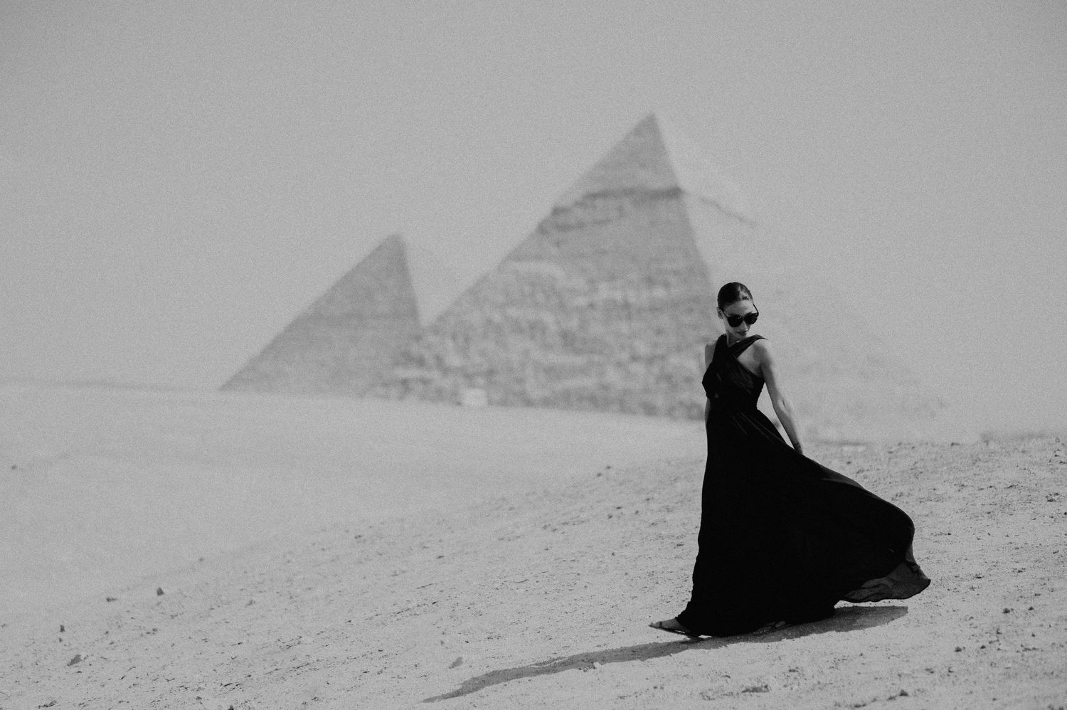 Historic black and white photograph showing someone sitting on sand in front of the Egyptian pyramids of Giza.
