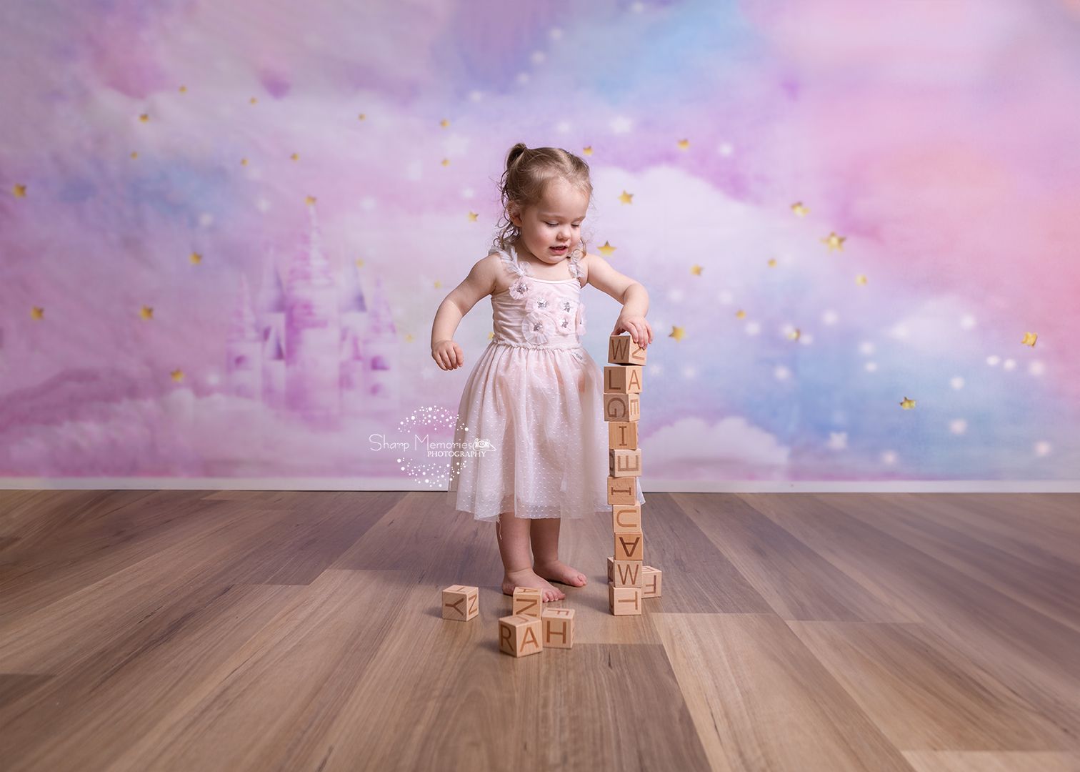Child in pink dress plays with wooden blocks against dreamy purple backdrop with sparkles and wooden floor.