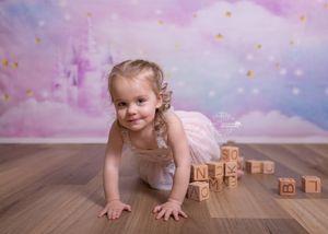 A child in a pink dress plays on a wooden floor with toy blocks against a dreamy pink and purple backdrop.