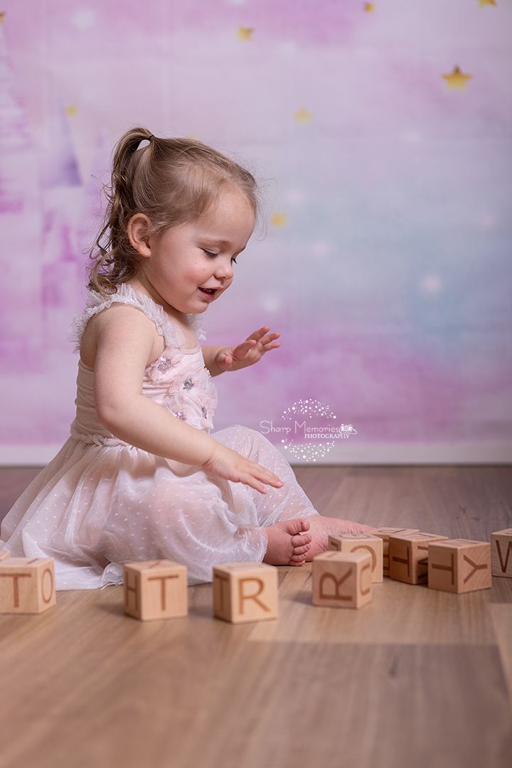 A dreamy portrait of a small child in a pink tulle dress playing with wooden letter blocks on hardwood flooring.