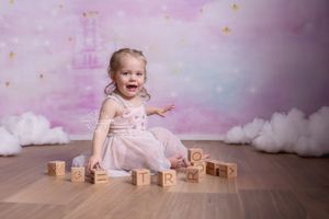 A toddler wearing a pink dress sits on a wooden floor playing with alphabet blocks against a dreamy pastel backdrop.