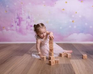 Child in pink dress playing with wooden alphabet blocks on hardwood floor against dreamy castle backdrop.