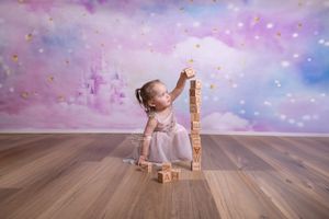 Small child in a pink dress stacks wooden alphabet blocks against magical purple and blue backdrop on hardwood floor.