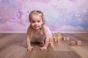 A young child in a pink dress crawls on hardwood floor with wooden alphabet blocks in a dreamy pink and purple setting.