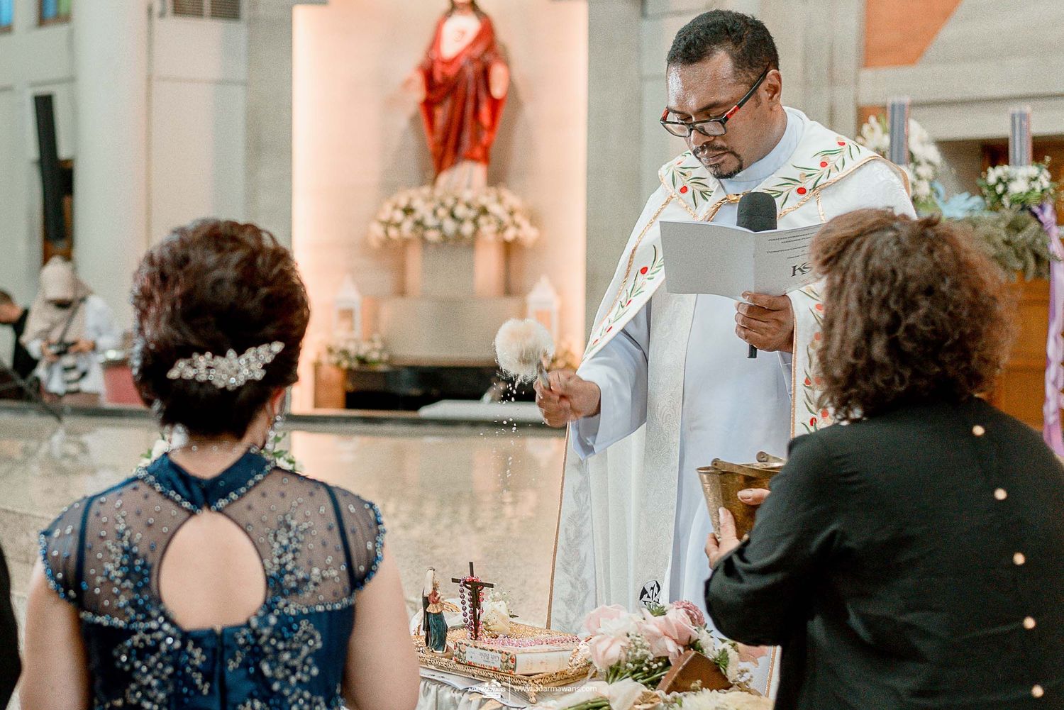 Church ceremony with priest presiding at decorated altar with religious statue in background.