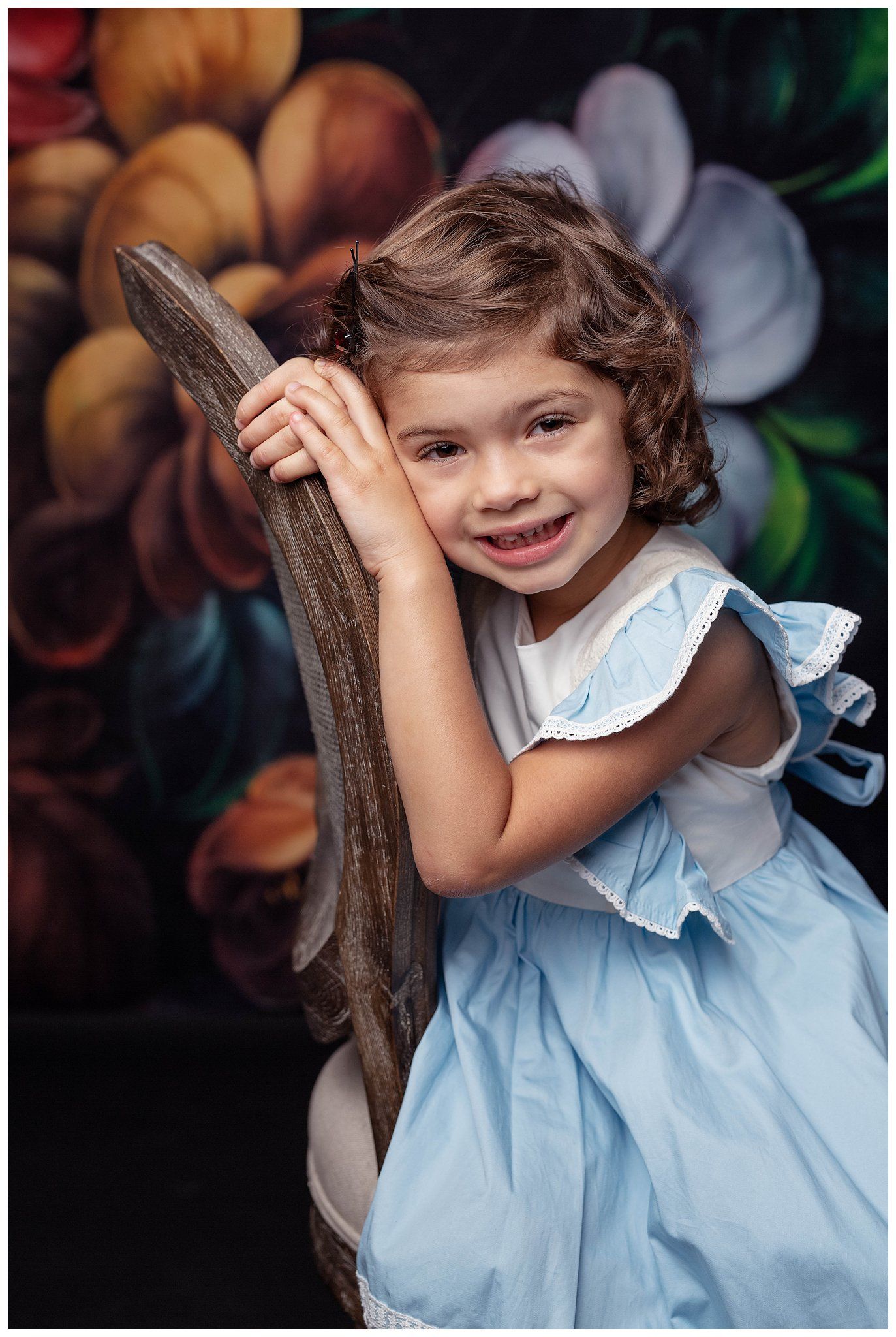 Smiling girl in light blue dress posing in front of floral backdrop during fine art child photography session in Austin