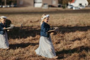 Dancing figures in blue denim tops and flowing white skirts twirl gracefully in a rustic field at sunset.