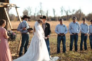 Outdoor wedding ceremony with attendees gathered under a wooden arch in a rustic countryside setting.