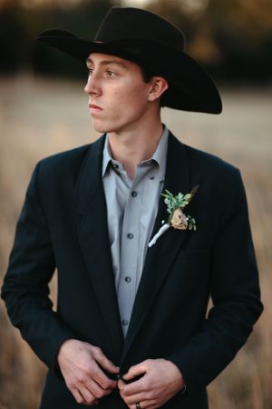 A groom in a black cowboy hat and suit with boutonniere poses against a blurred background.