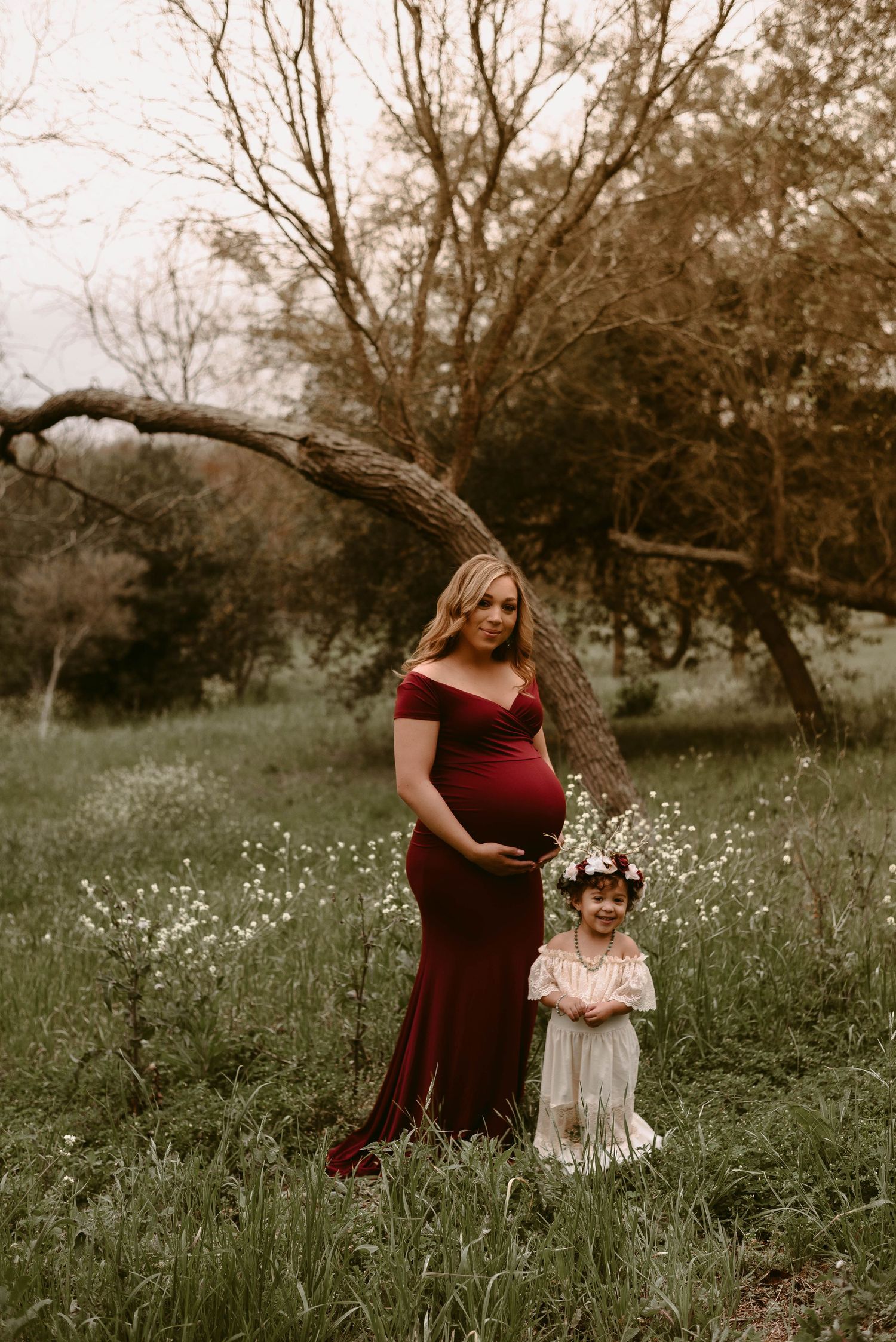 A maternity portrait under a curved tree branch in a wildflower meadow.