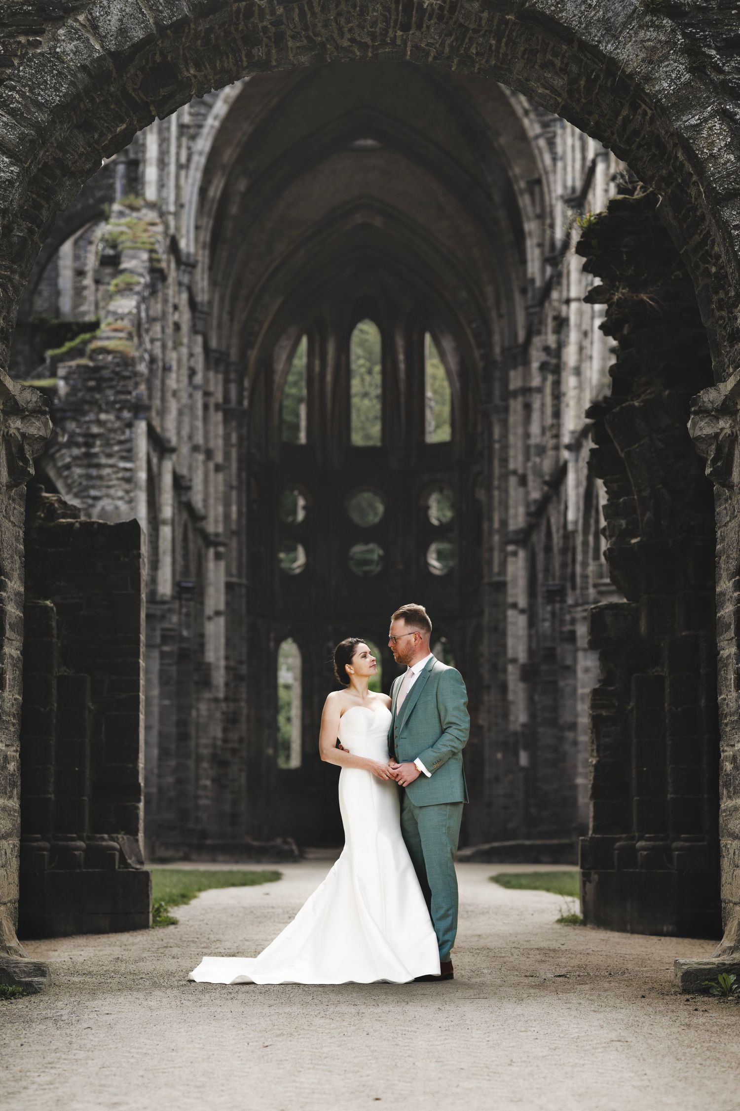 Wedding couple poses in elegant attire under gothic stone archway of historic ruins.