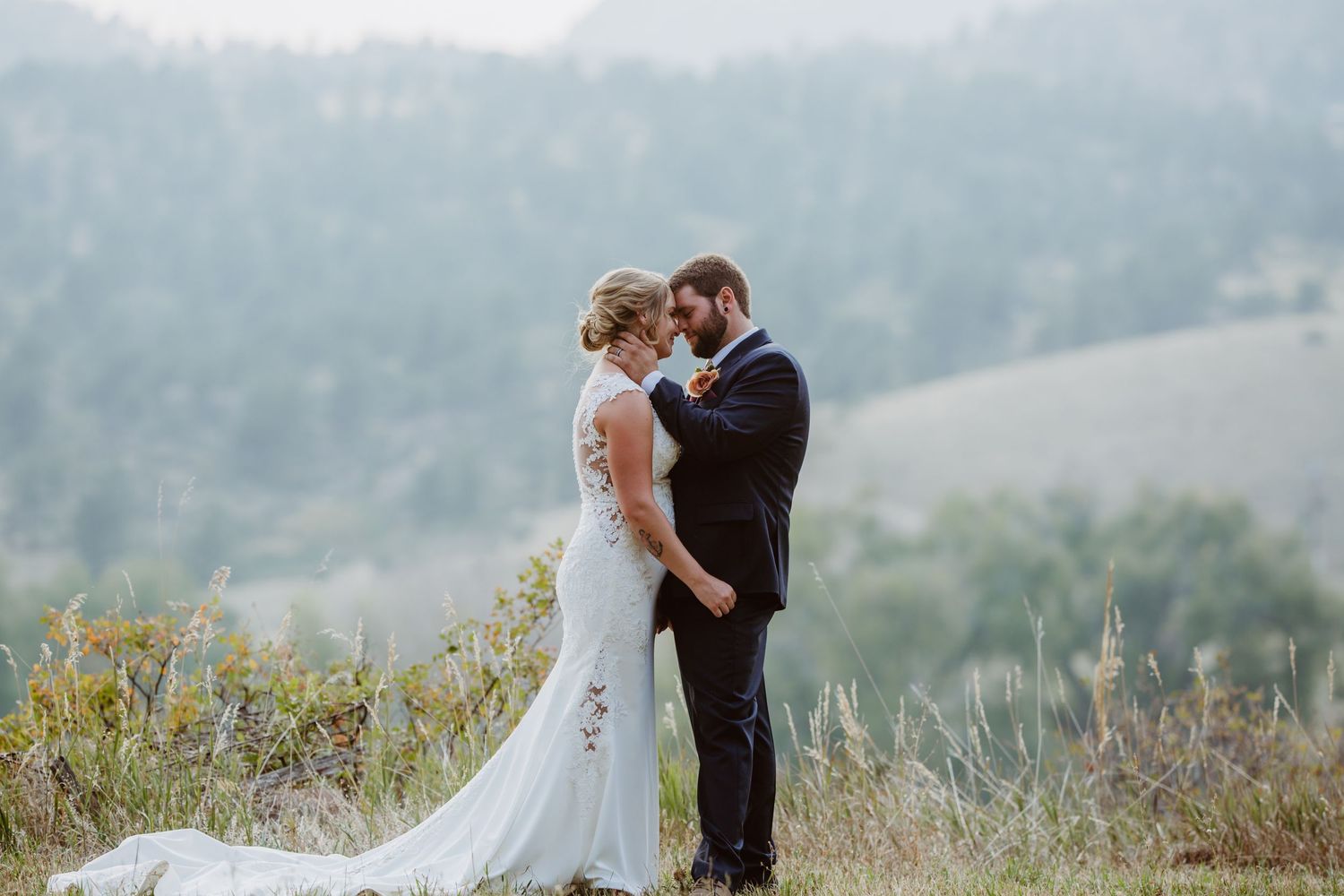 A romantic wedding couple embraces on a misty hillside surrounded by wildflowers and mountains.
