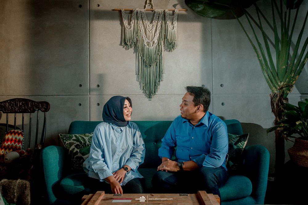 Couple having conversation while seated on teal couch with macrame wall hanging in background.