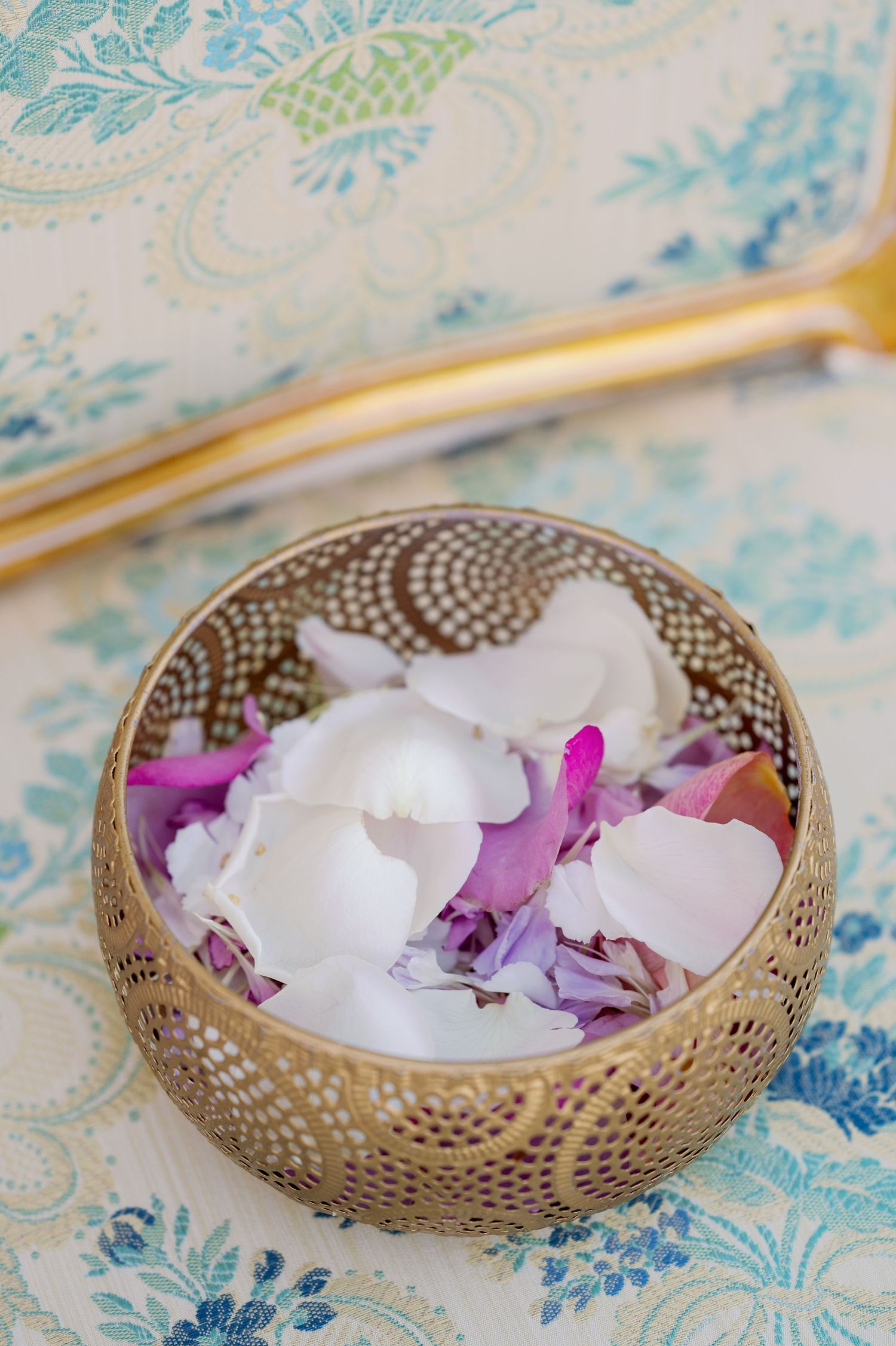 Delicate white and purple flower petals in an ornate gold bowl on blue patterned fabric.