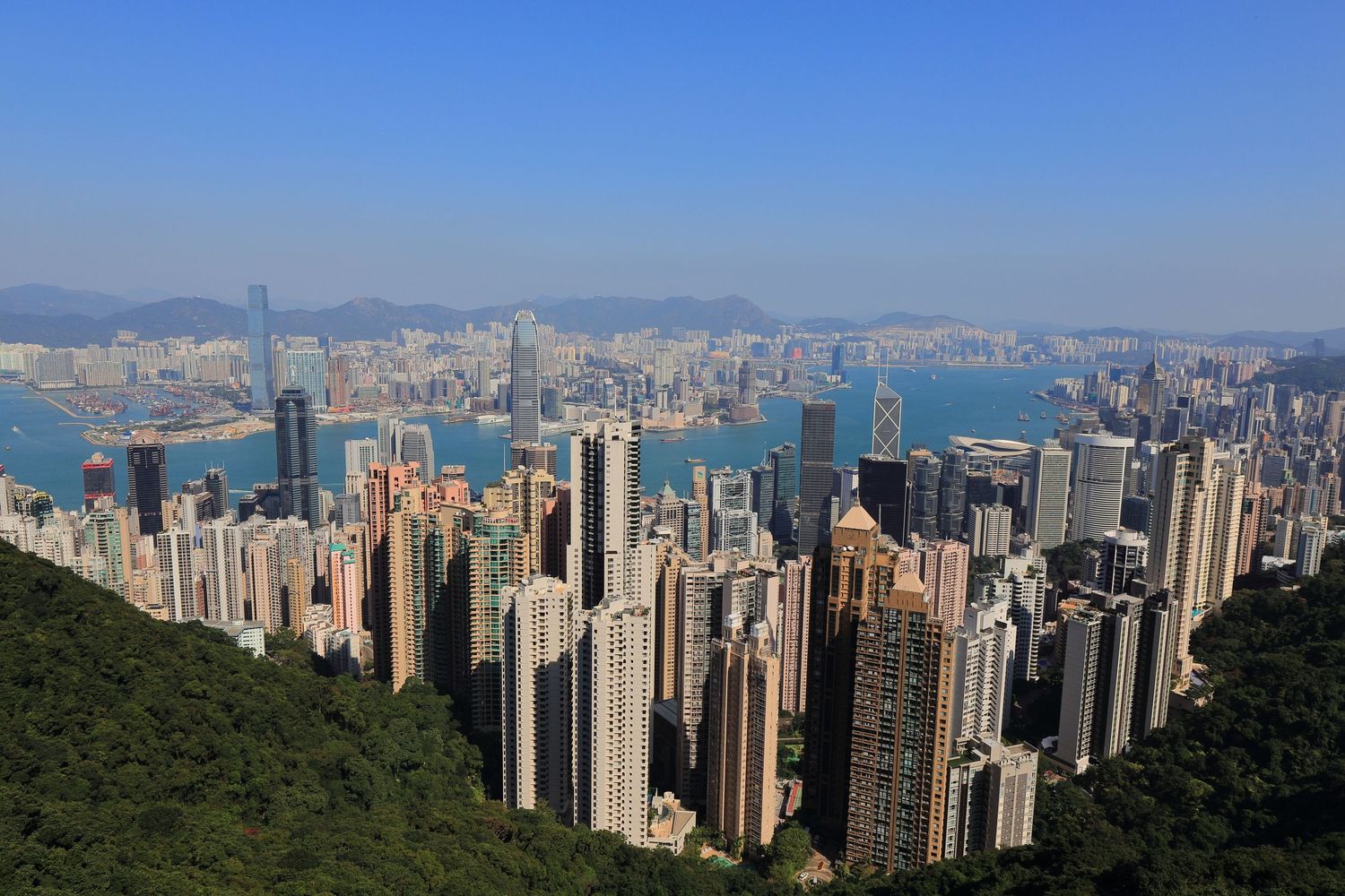Aerial view of Hong Kong's iconic skyline with Victoria Harbor and mountains in background.