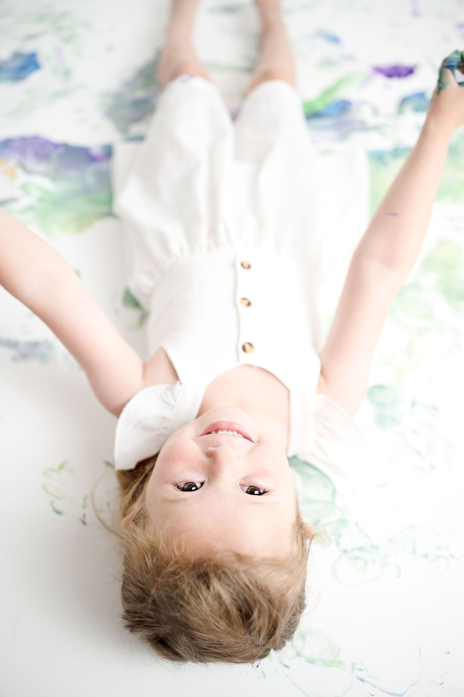 Child lying on white floor surrounded by colorful paint splatters.