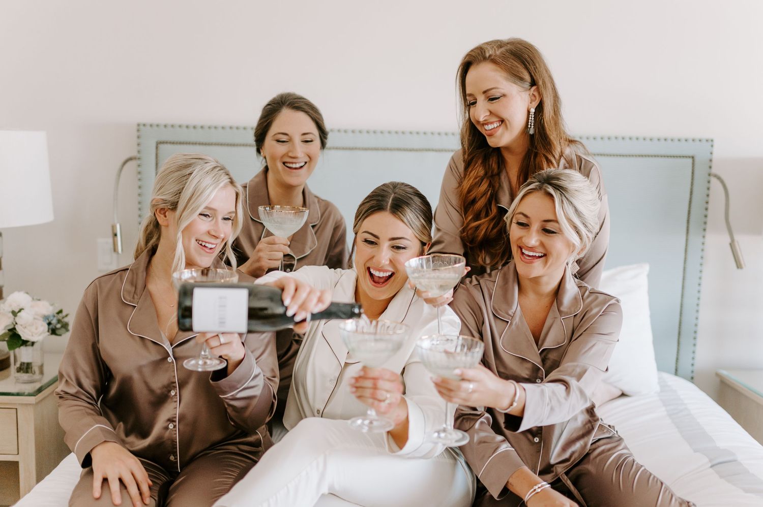 Group in matching robes share laughs while looking at phones on bed during wedding preparations.