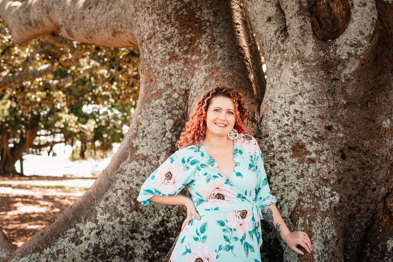 A person in turquoise floral dress poses by large tree trunk.