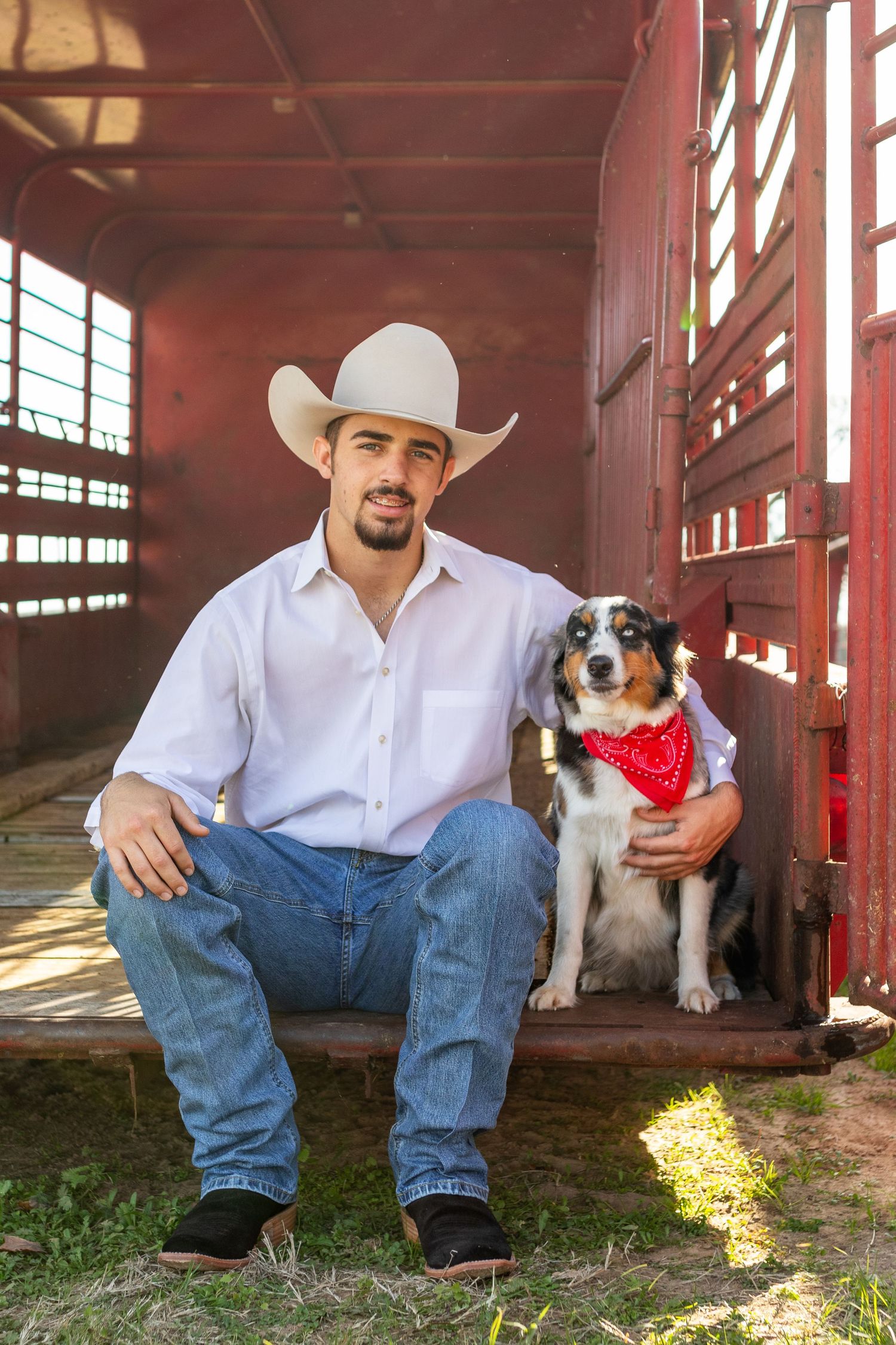 Cowboy in white hat sits with Australian Shepherd dog wearing red bandana at livestock trailer entrance.