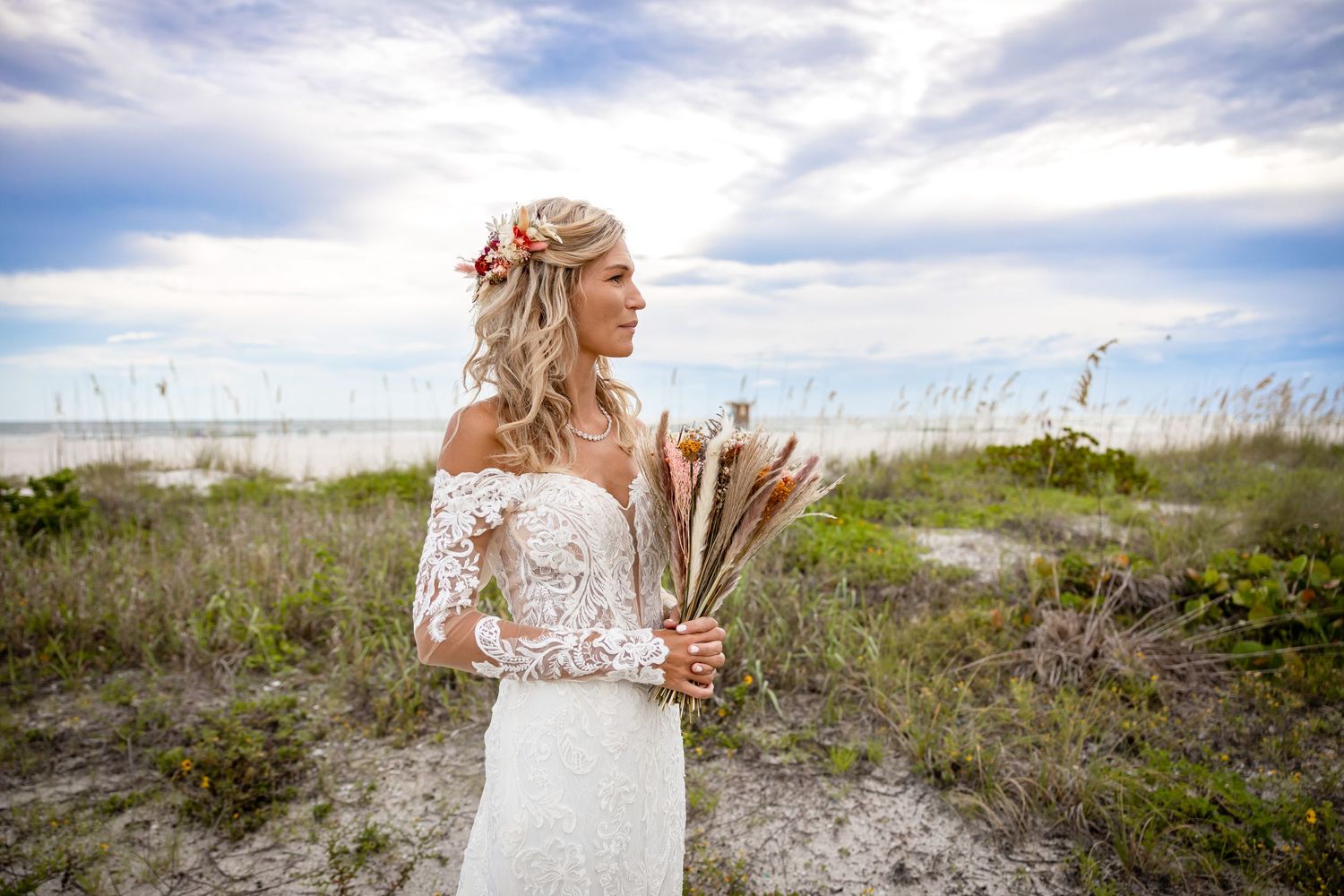 Bride in off-shoulder lace gown holds dried floral bouquet among beach dunes under cloudy sky.