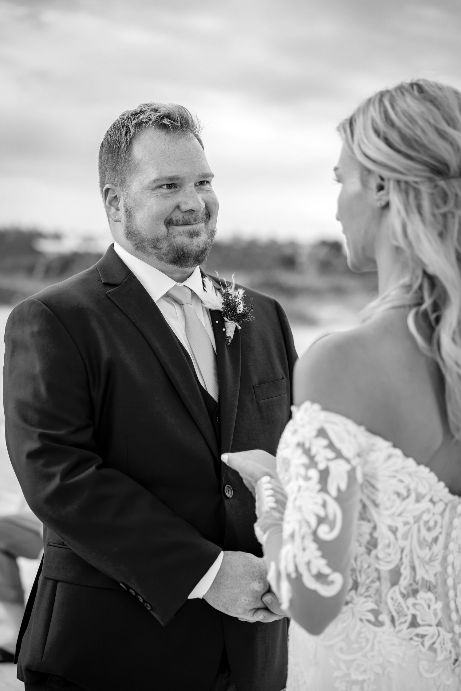 Black and white portrait of groom smiling tenderly at bride during beach wedding ceremony.