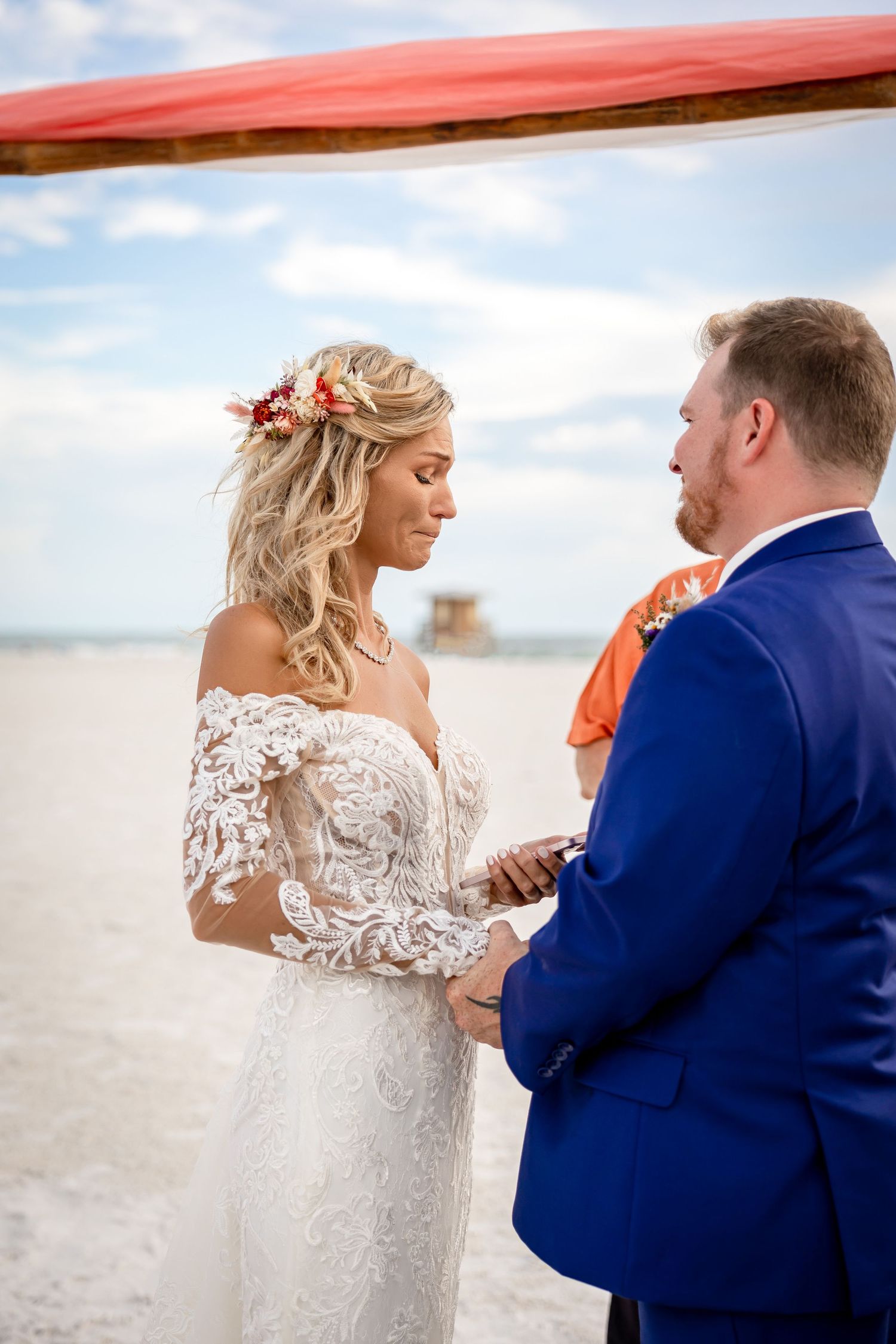 Bride in lace gown exchanges vows with groom in blue suit under coral arch at beach ceremony.