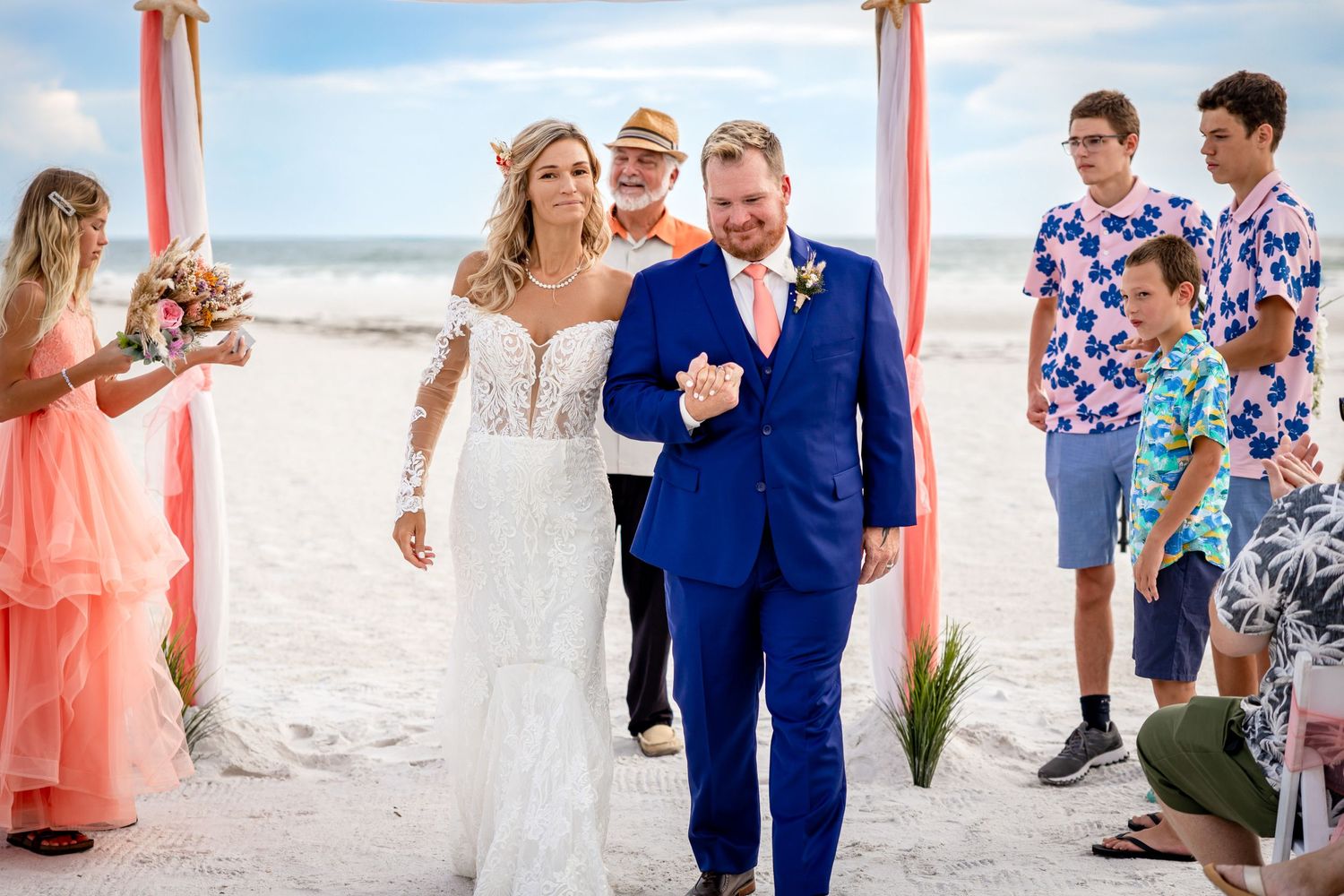 Newlyweds walk down sandy beach aisle under coral arch, surrounded by smiling wedding guests.