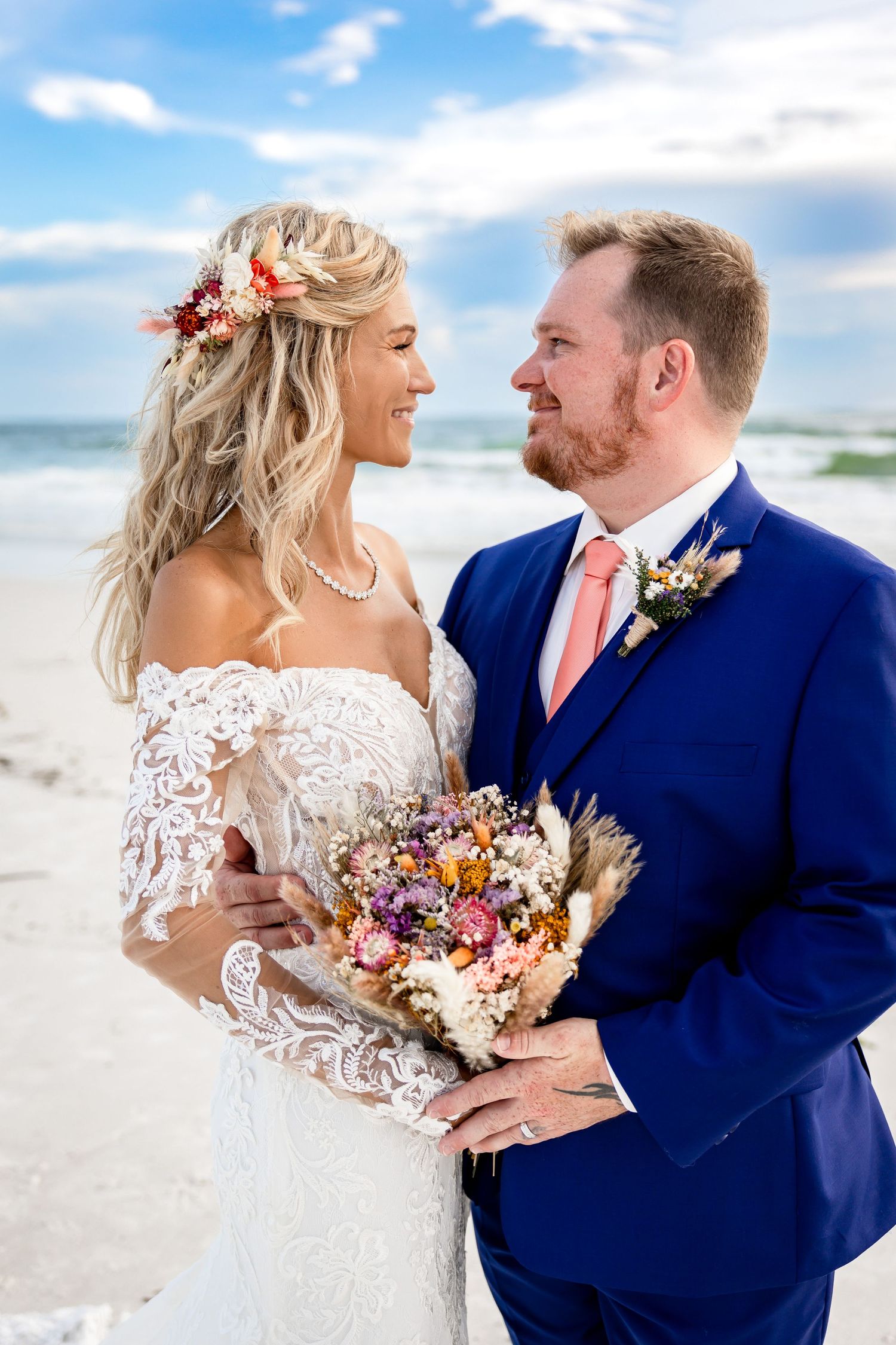 Smiling newlyweds embrace on beach, bride holding colorful dried bouquet, groom in navy suit.
