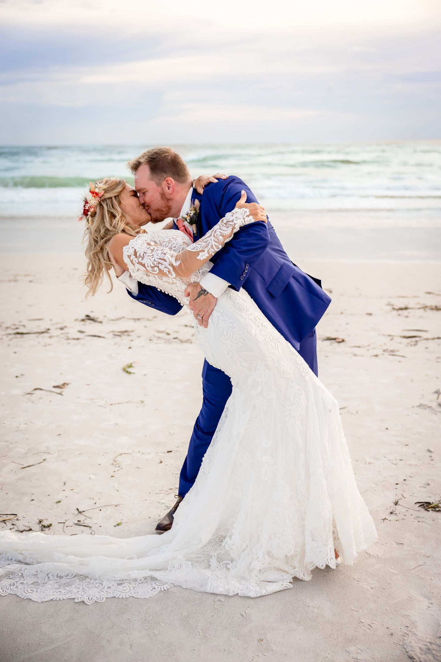 Groom dips bride for a kiss on the sandy beach with ocean waves in the background.