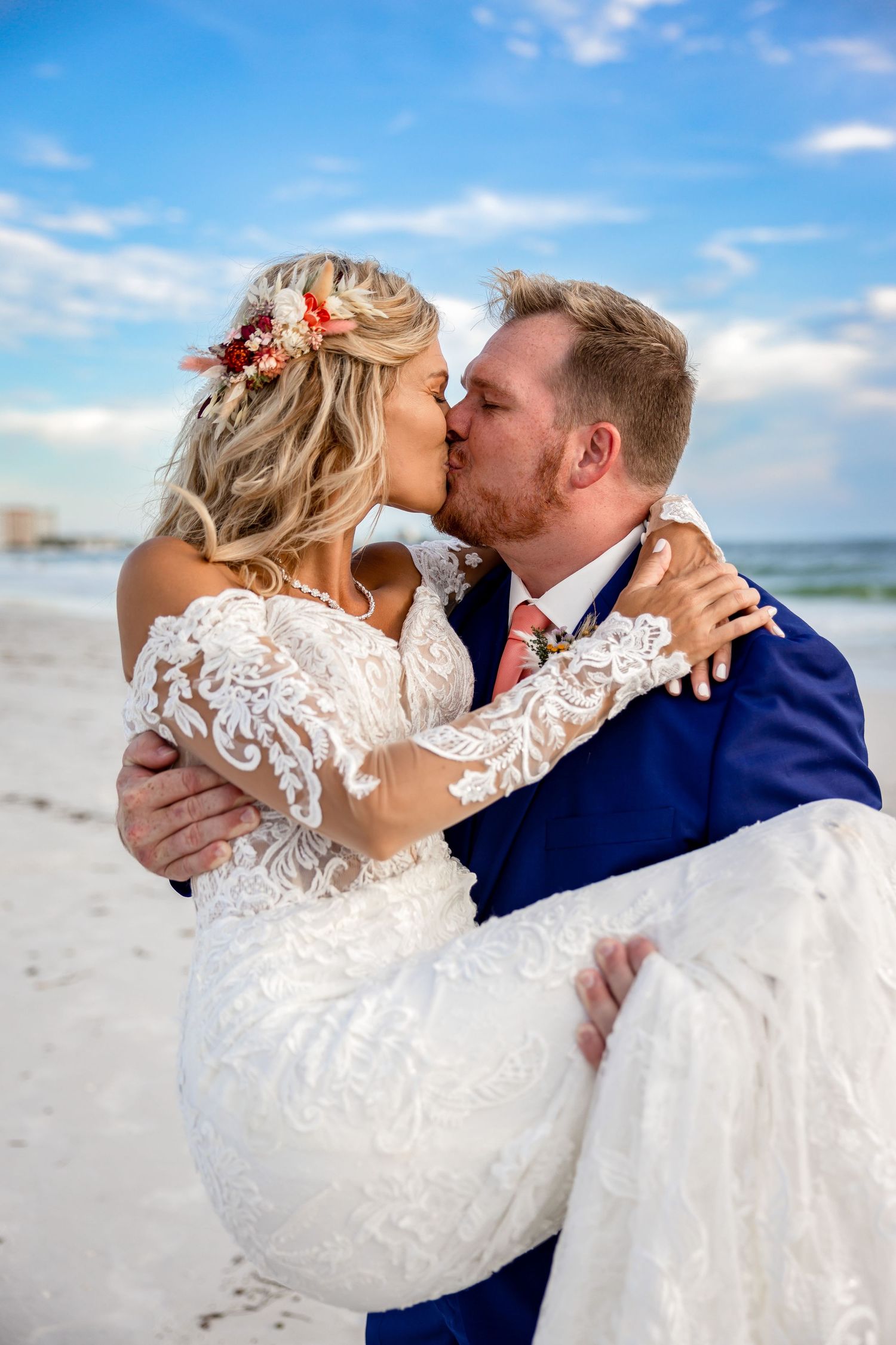 Groom in navy suit carries and kisses bride in lace gown on sunny beach after wedding.