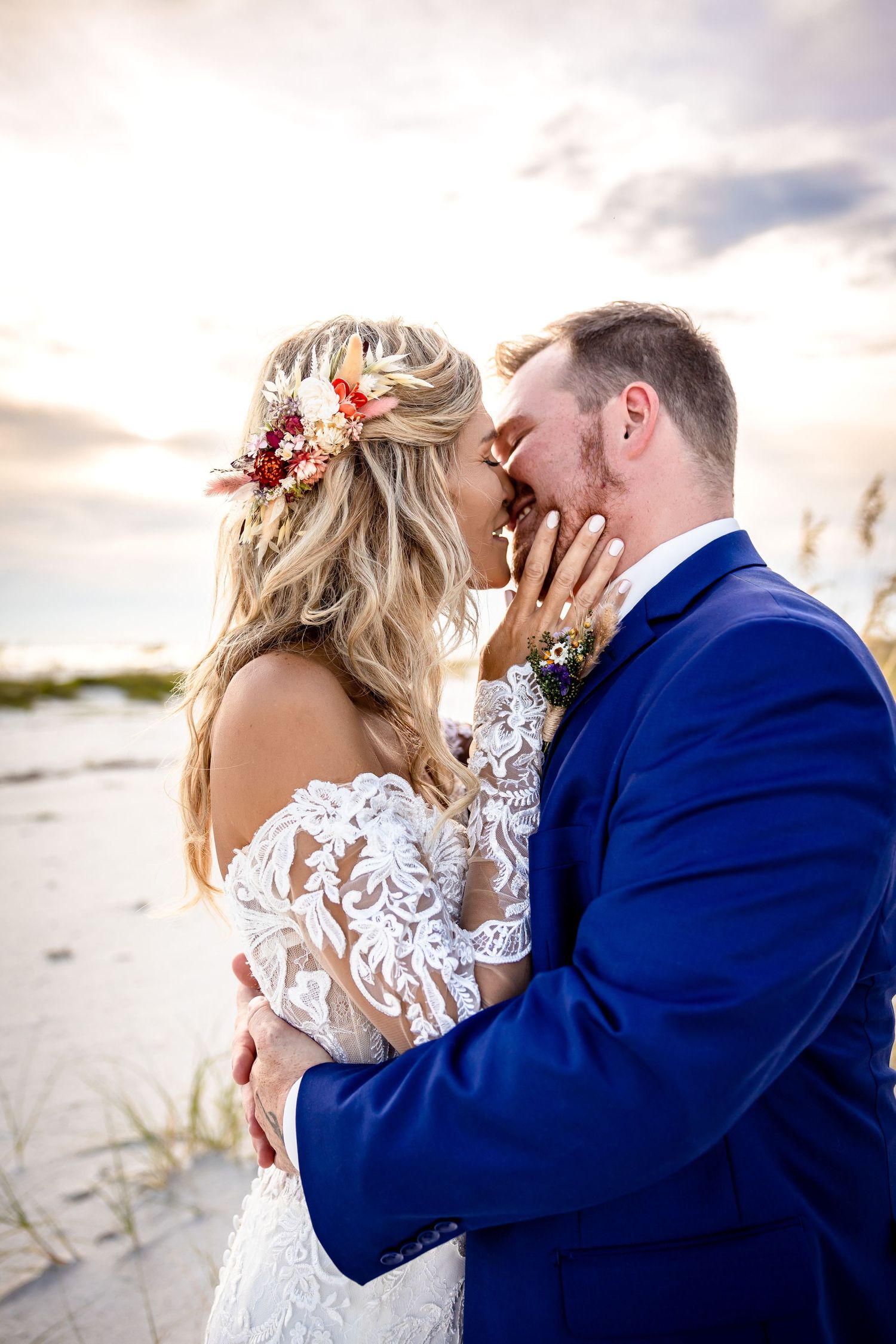 Couple shares a tender sunset kiss on the beach, bride in lace gown with floral hair crown.