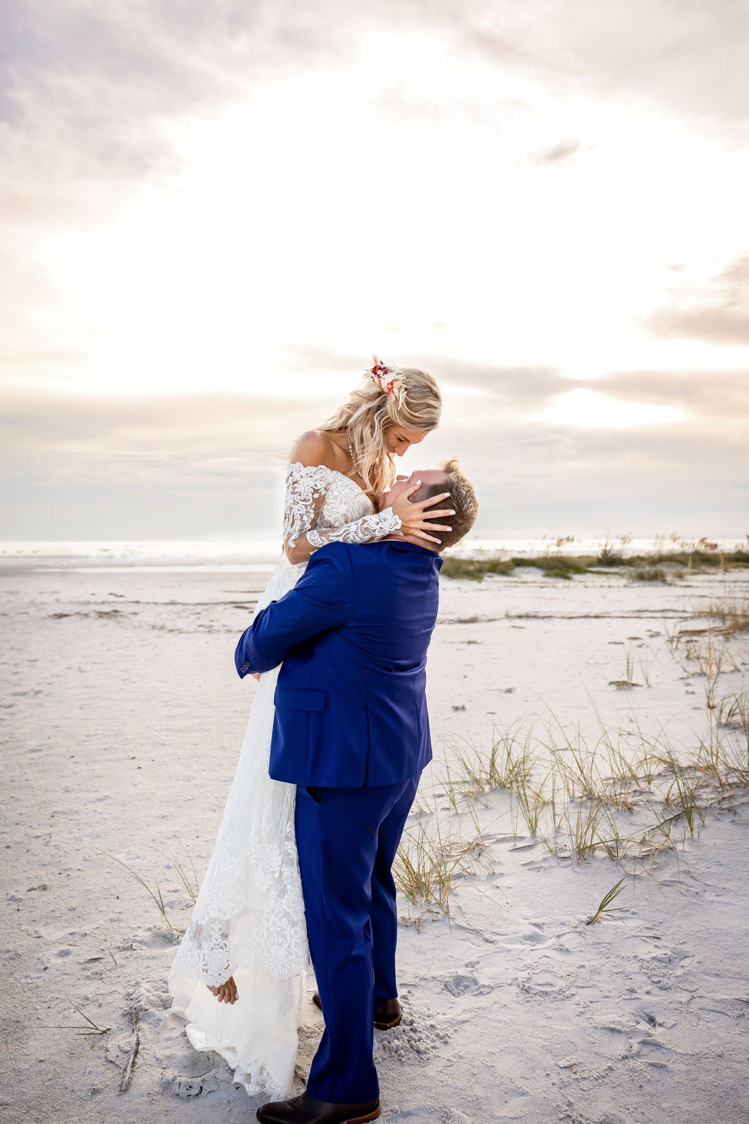 Bride and groom share a romantic kiss on a sandy beach at sunset, groom in blue suit lifting bride in lace wedding gown.