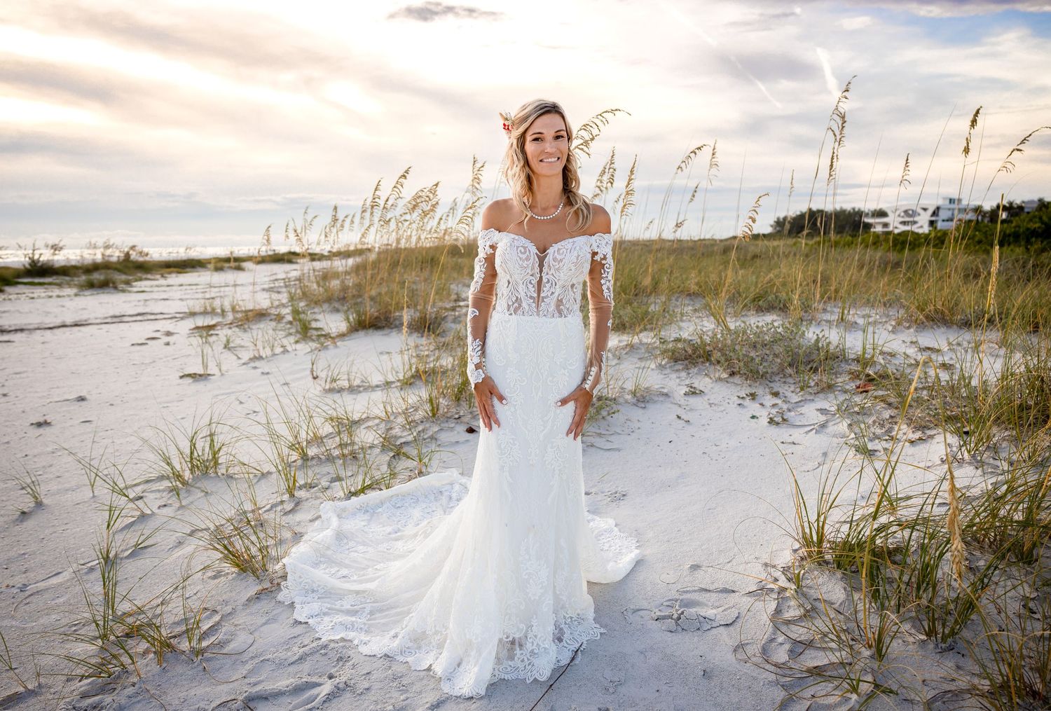 Bride poses alone on a sunny beach in an elegant off-shoulder lace mermaid wedding dress with sea oats in the background.