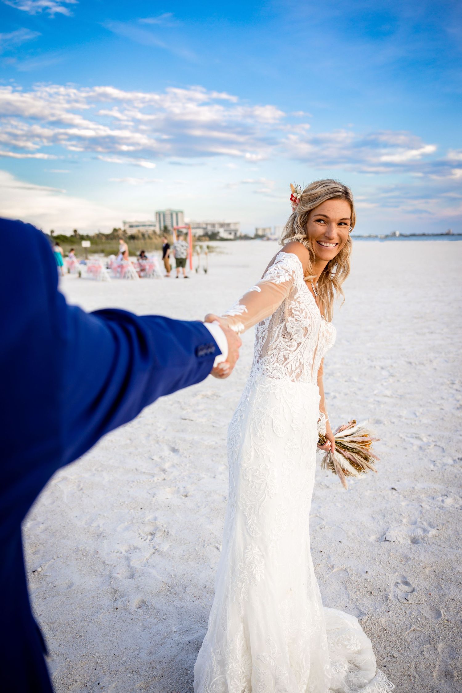 Smiling bride in lace wedding gown holds groom's hand on white sand beach while carrying a boho dried flower bouquet.