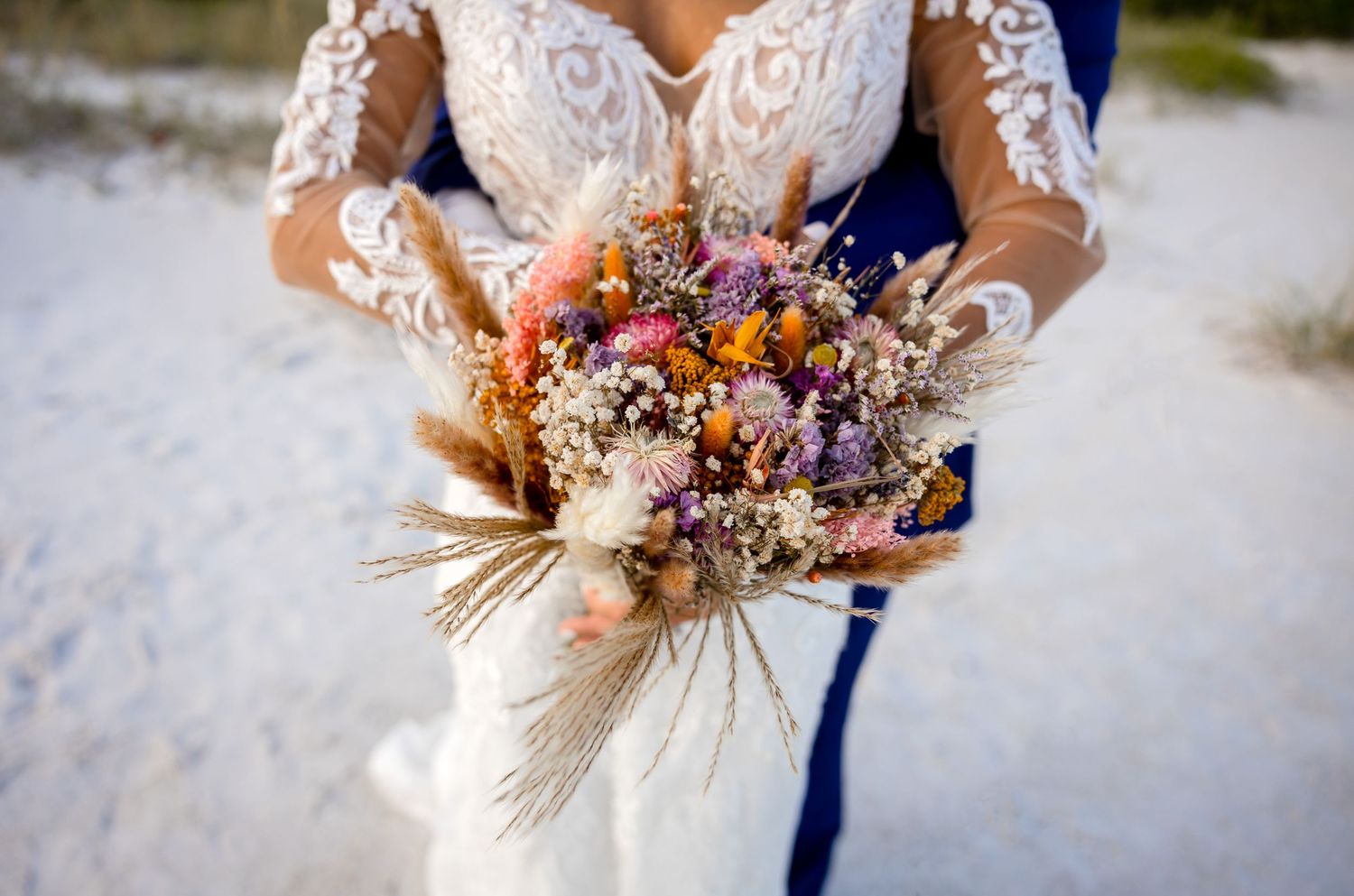 Couple holds a colorful boho dried flower bridal bouquet with pampas grass and wildflowers on a sandy beach backdrop.