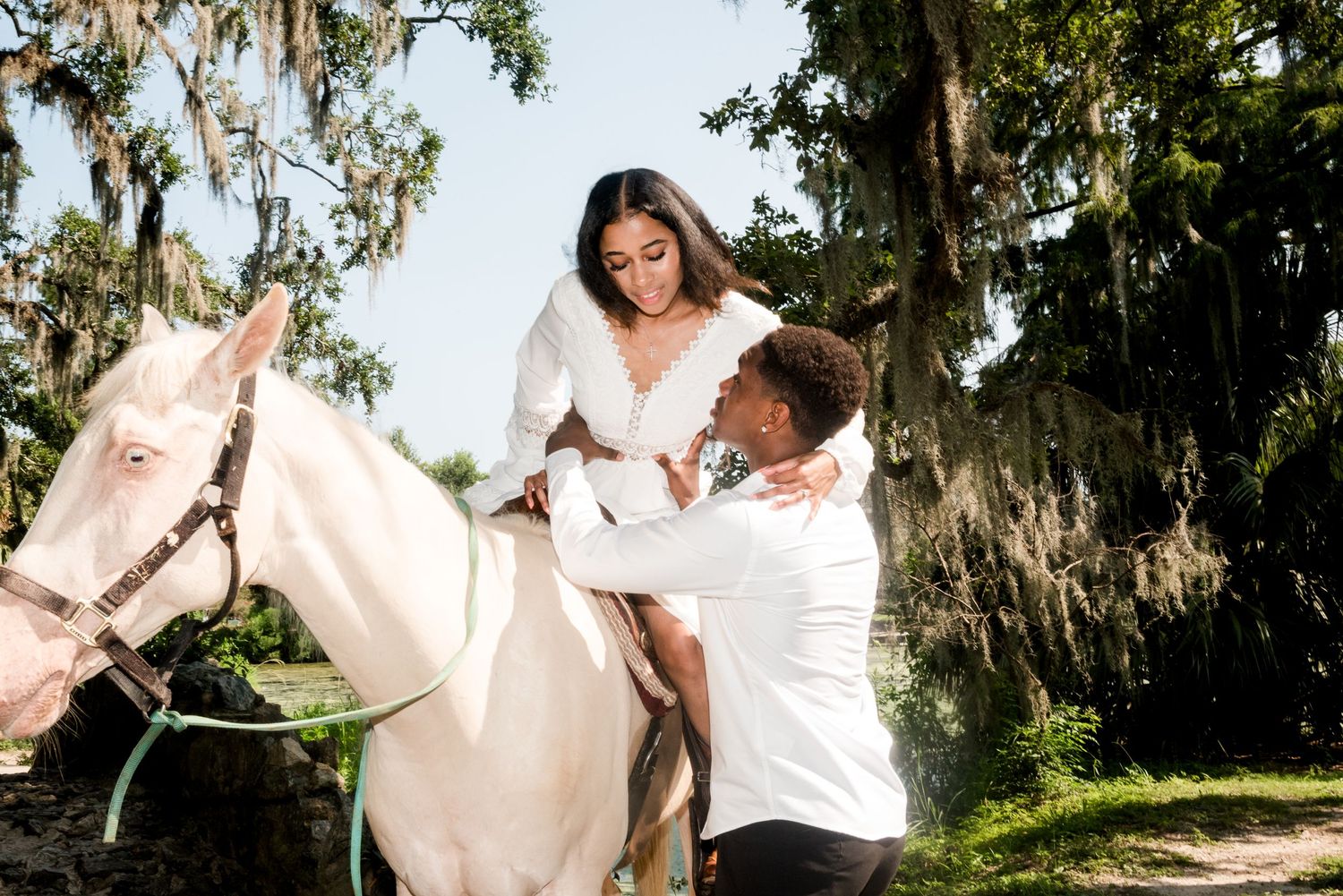 Couple in white wedding attire posing with white horse under moss-draped trees.