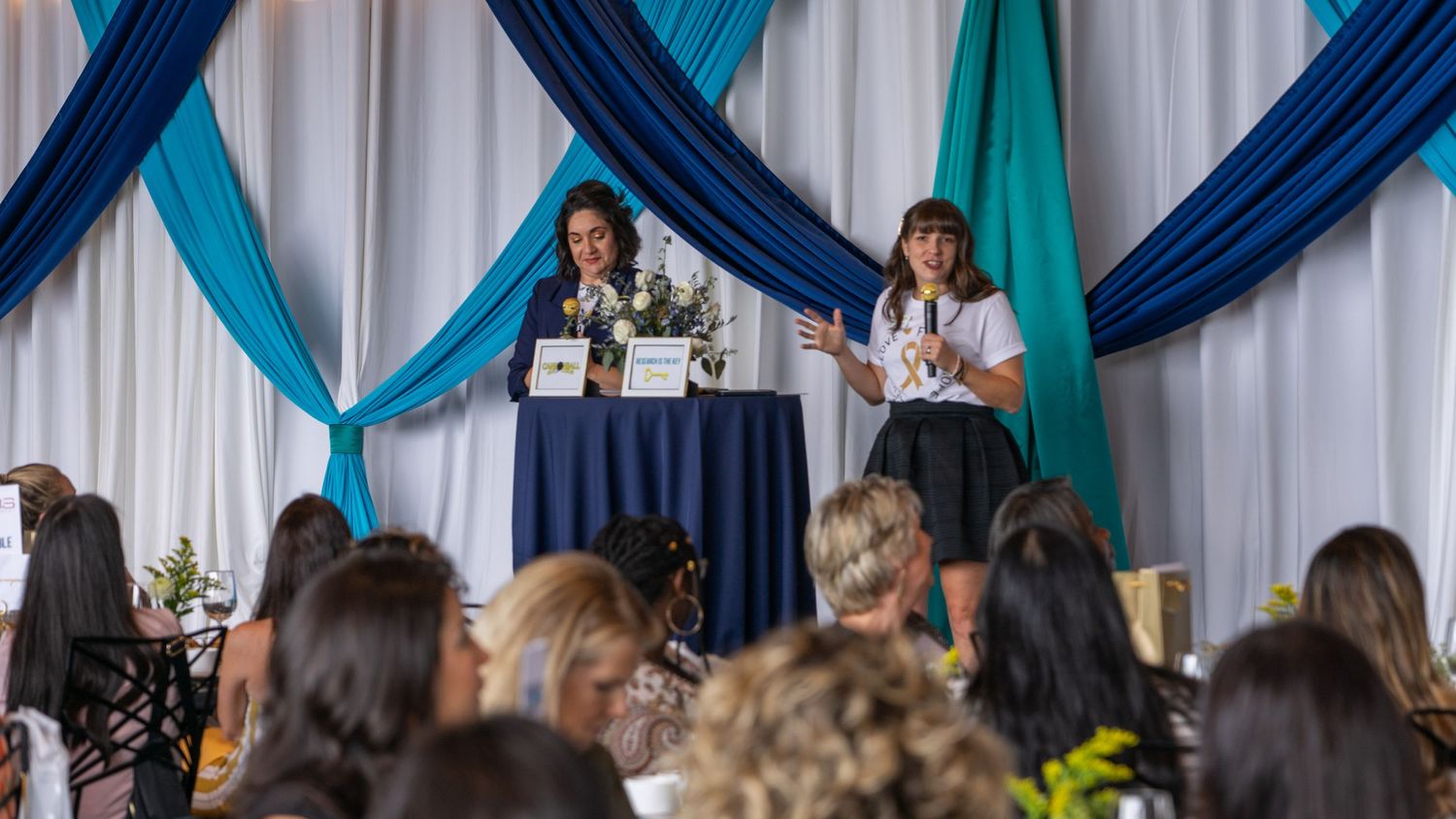 Speakers presenting on stage with blue and teal draped backdrop at a women's conference.