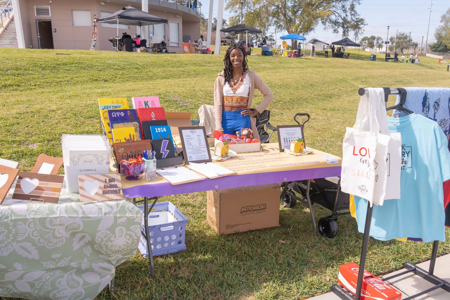 Outdoor vendor booth displaying books, clothing and merchandise on a sunny day.
