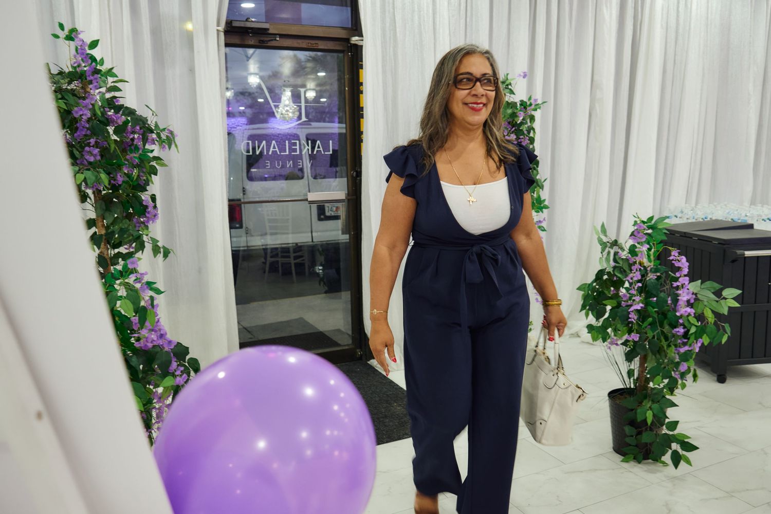 Woman in navy jumpsuit stands near purple balloon and floral decorations.