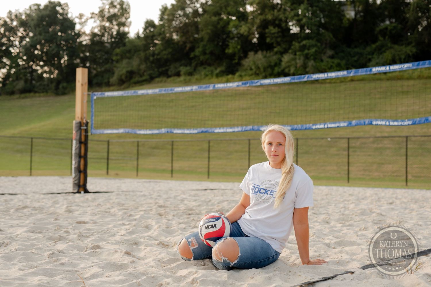 Athlete relaxes on sandy volleyball court with volleyball and ripped jeans.