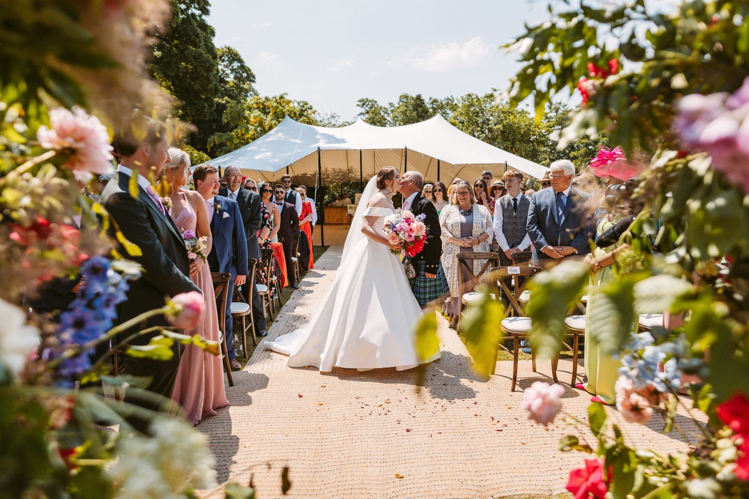 Bride arriving with Father dad walks beautiful daughter up the aisle surrounded by bold colourful flowers summers day