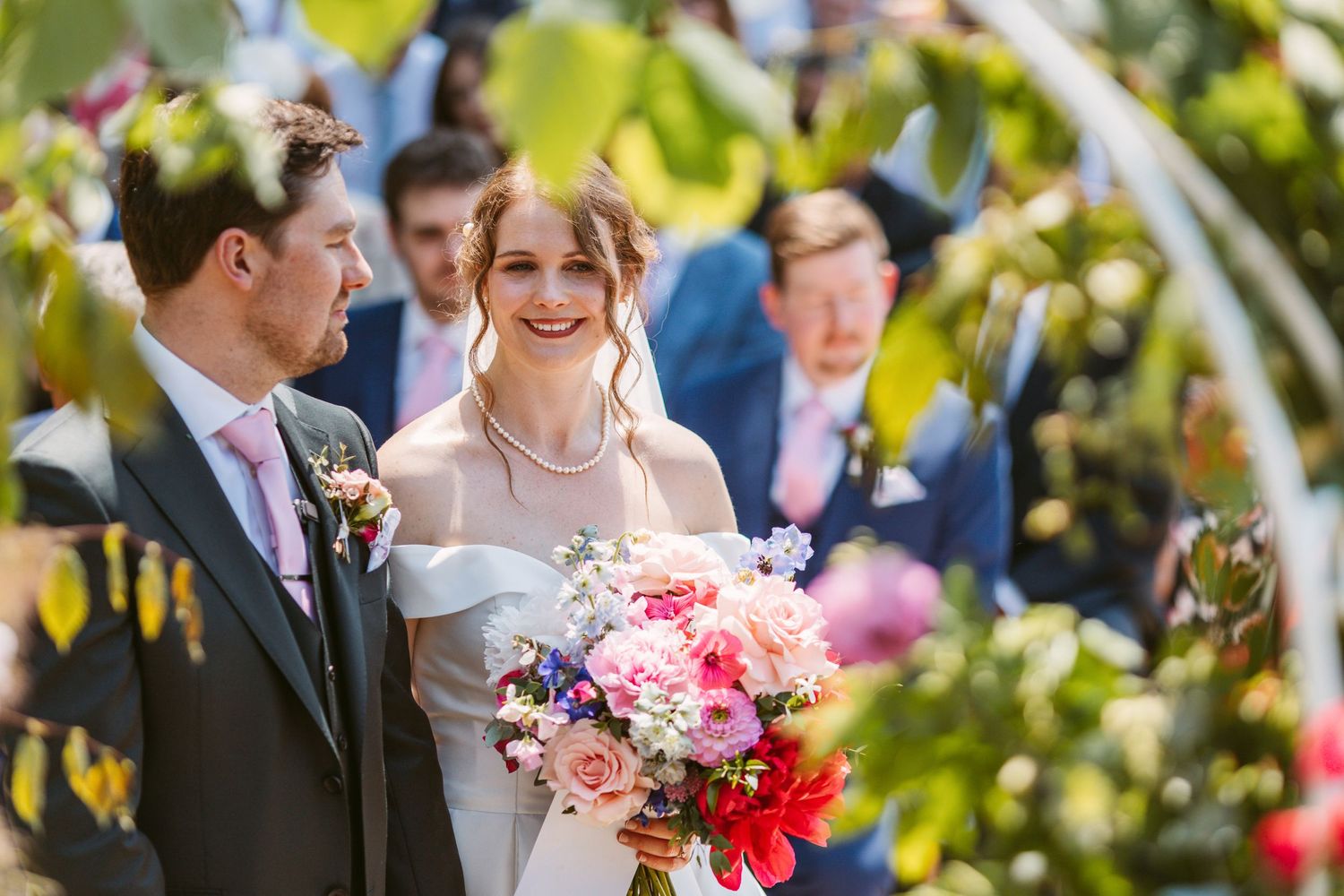 Bride arriving at top of aisle and greeting groom surrounded by bold colourful flowers summers day